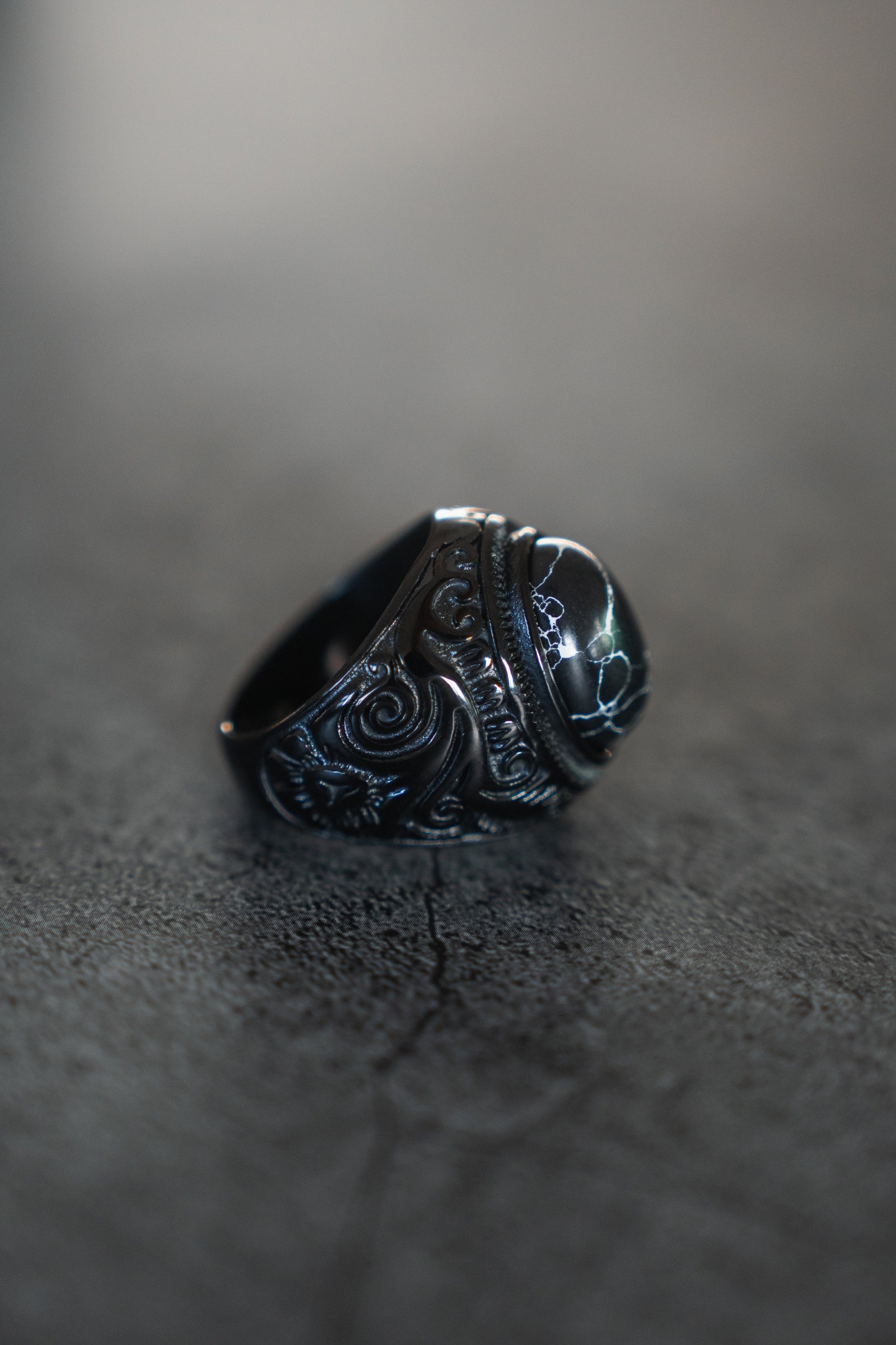 Displayed on a dark, textured backdrop, The Dark Storm Ring by Relentless Betrayal features intricate swirling designs on its silver band. The lighting accentuates the engraved details and the glossy centerpiece of this mystical artifact.
