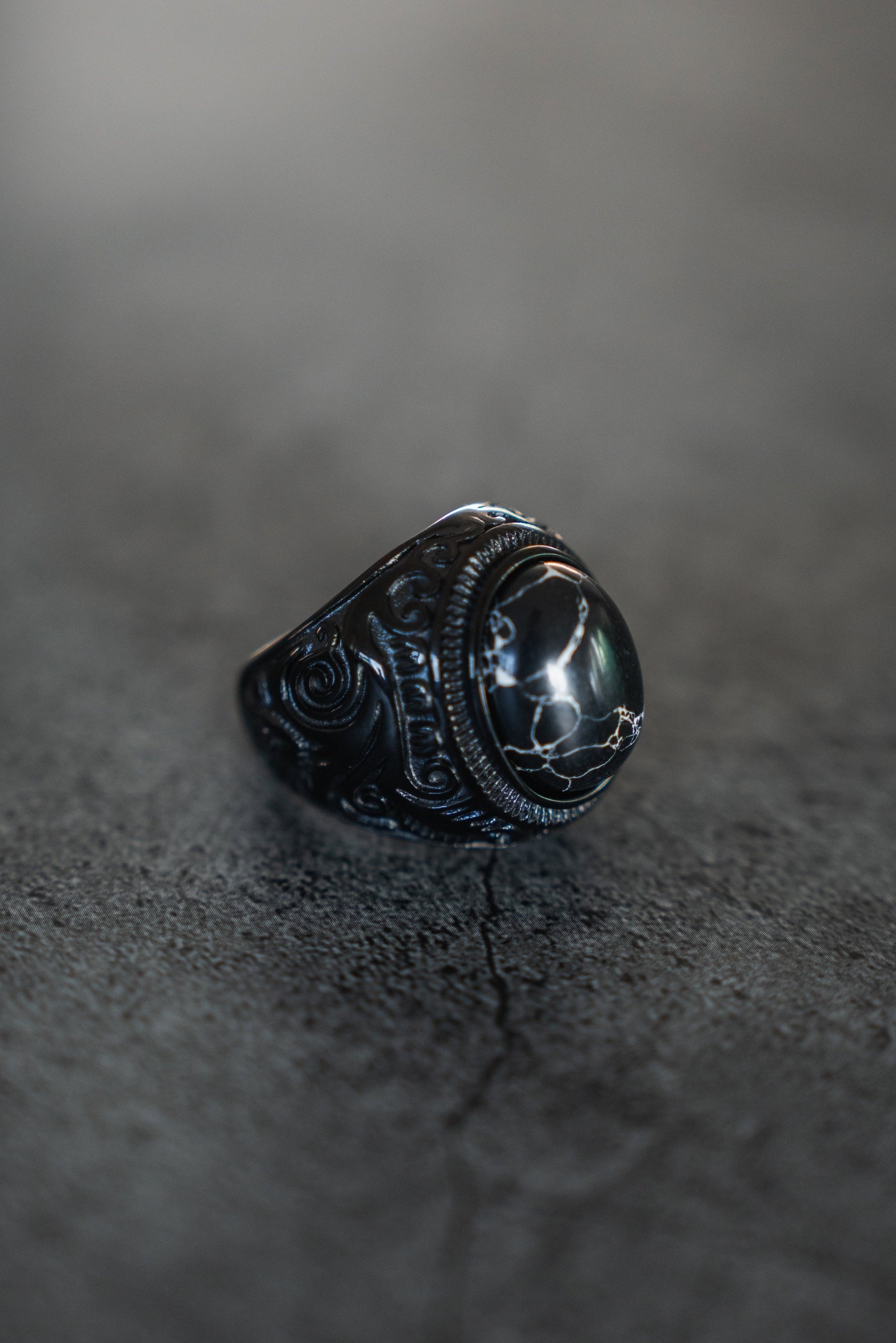 The Dark Storm Ring by Relentless Betrayal is a mystical artifact crafted from stainless steel with intricate carvings on a textured gray surface, featuring a dark stone center adorned with white crack-like patterns, exuding ornate vintage charm.