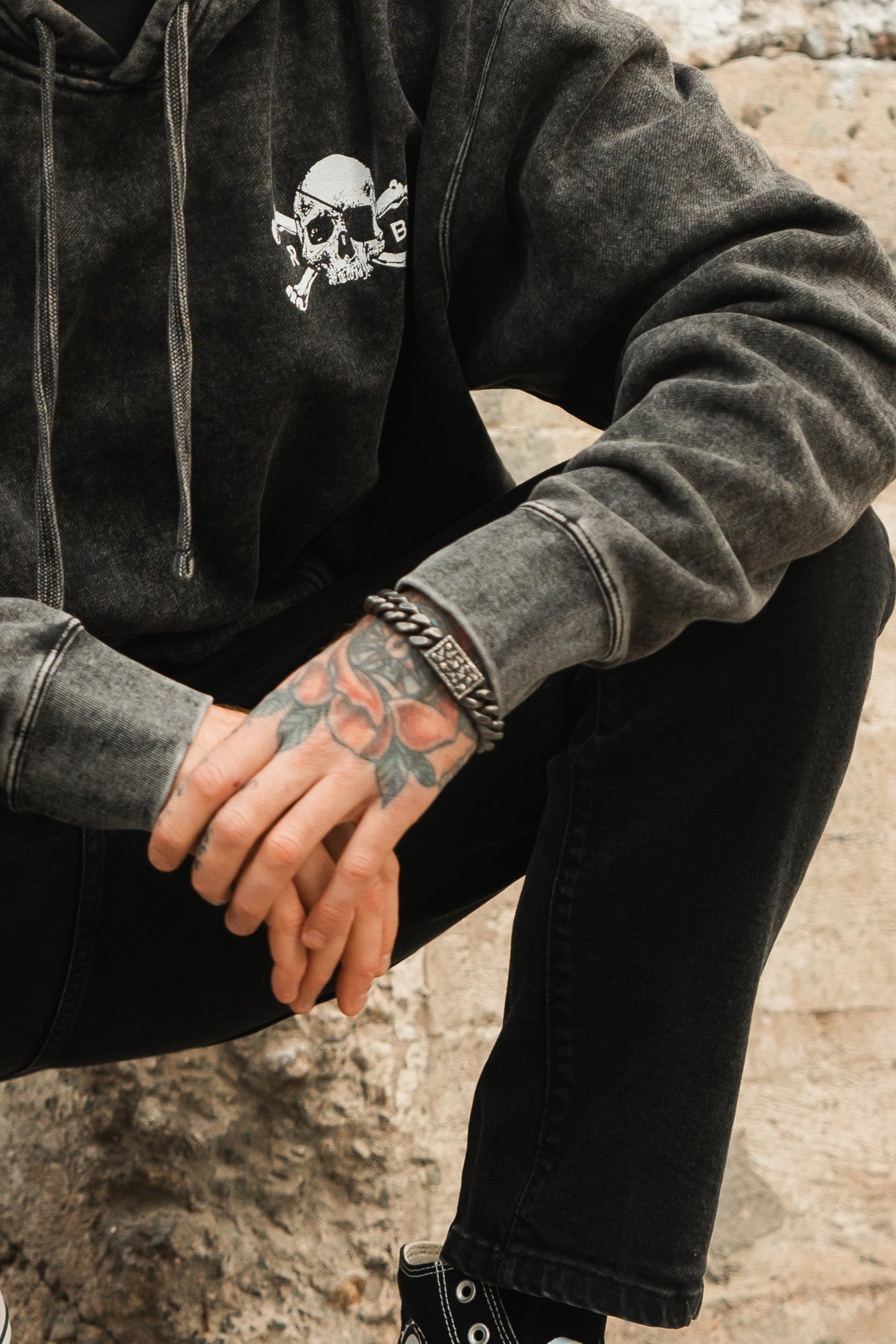 A person in a dark hoodie featuring a skull and crossbones graphic sits with hands clasped, showcasing a rose tattoo and the Tell No Tales Bracelet by Relentless Betrayal. Wearing black jeans and sneakers, they relax on a stone surface.