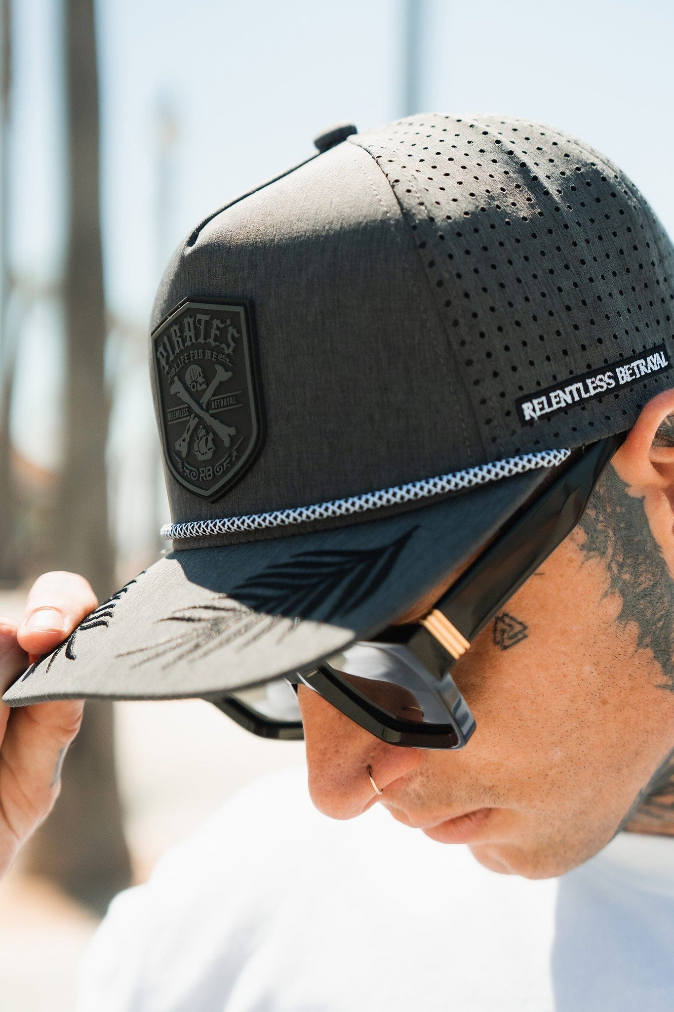 Wearing a Pirates Life Grey Snapback from Relentless Betrayal, featuring a PVC rubber patch logo and Relentless Betrayal along the side, a person pairs it with large sunglasses. The water-resistant hat stands out against blurred trees under the bright sun.
