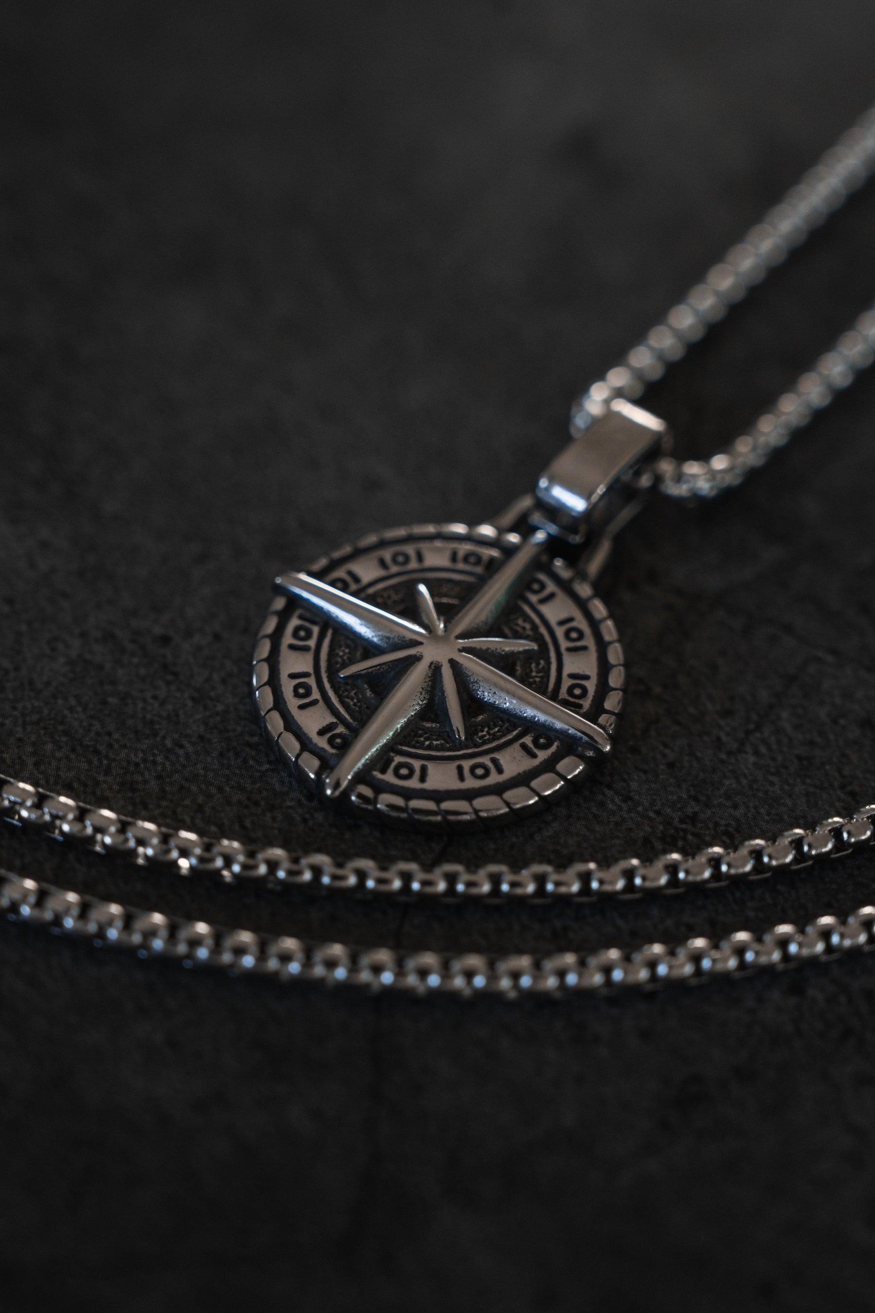 The Relentless Betrayal Polaris Necklace features a metallic compass design with sparkling gemstones. Its two layered chains enhance the intricate details, allowing the pendant to stand out beautifully against a dark background.