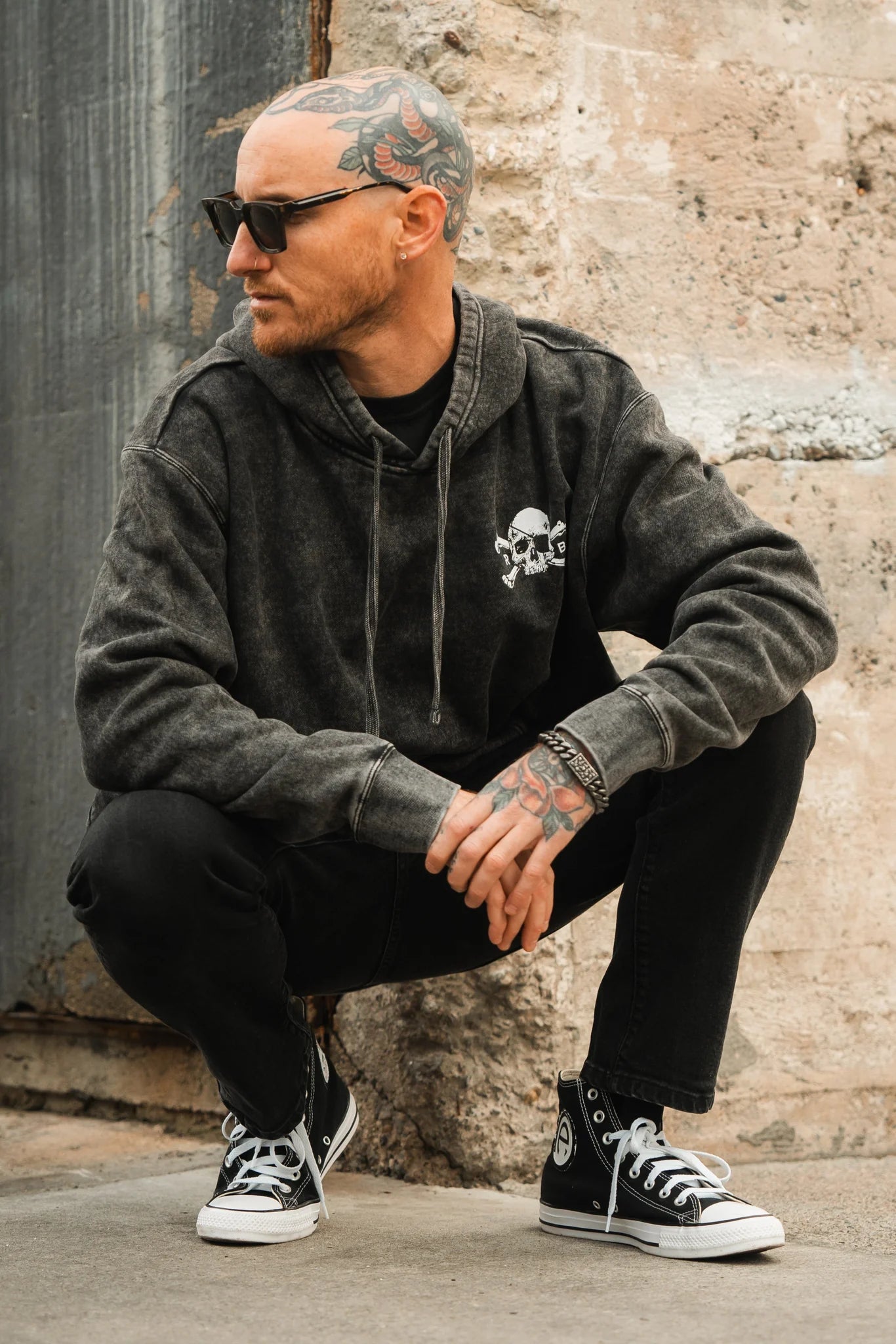A man squats outdoors, sunglasses shielding his eyes, in a Relentless Betrayal Tell No Tales Premium Hoodie featuring a skull design, black jeans, and high-tops. Tattoos adorn his head and hands as he leans against a stone wall, glancing sideways.