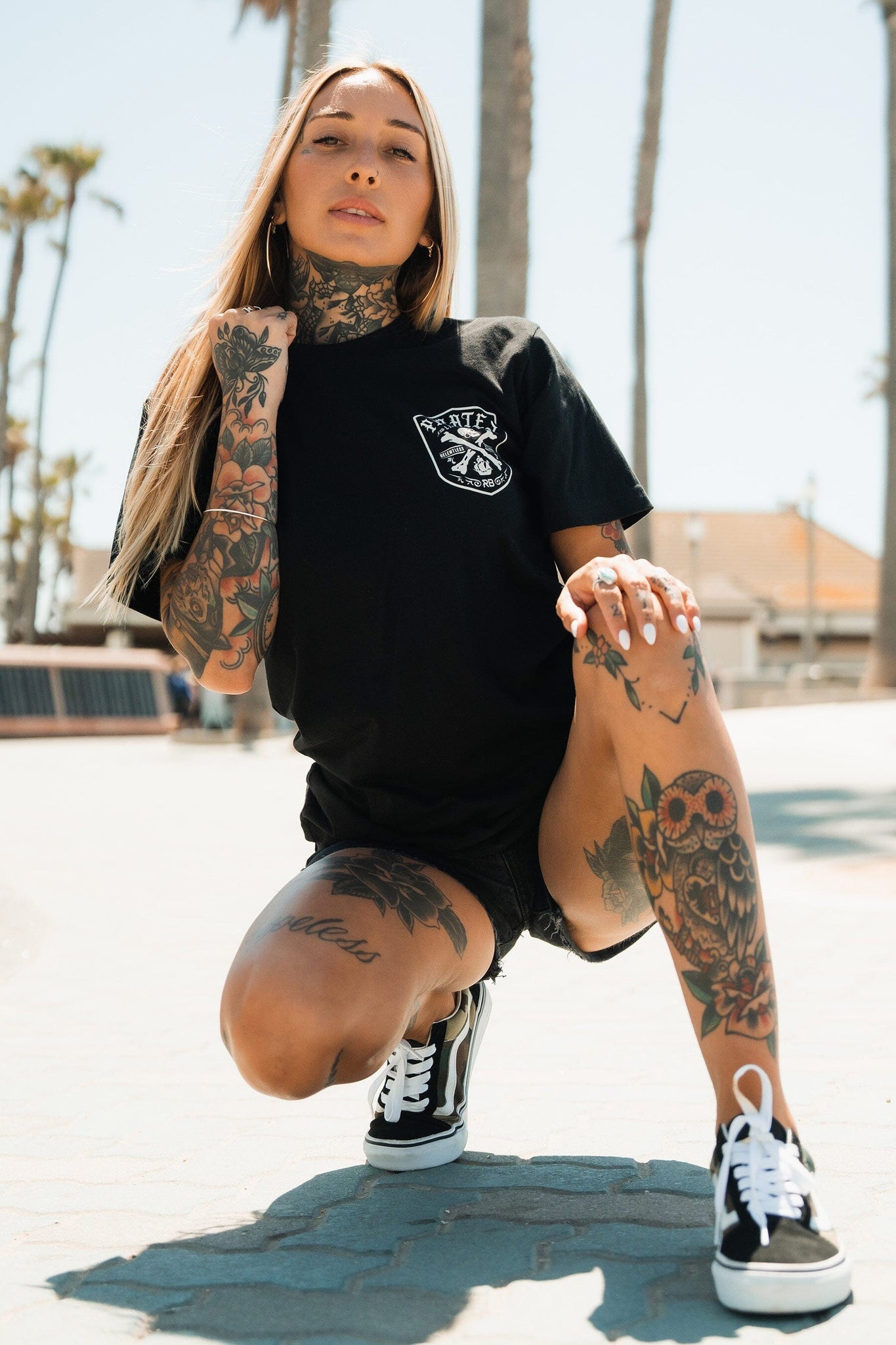 A confident person with long hair kneels amidst swaying palm trees, donning extensive tattoos and the Pirates Life Black T-Shirt by Relentless Betrayal. They pair it with black shorts and sneakers against a boardwalk backdrop, embodying an adventurous spirit.