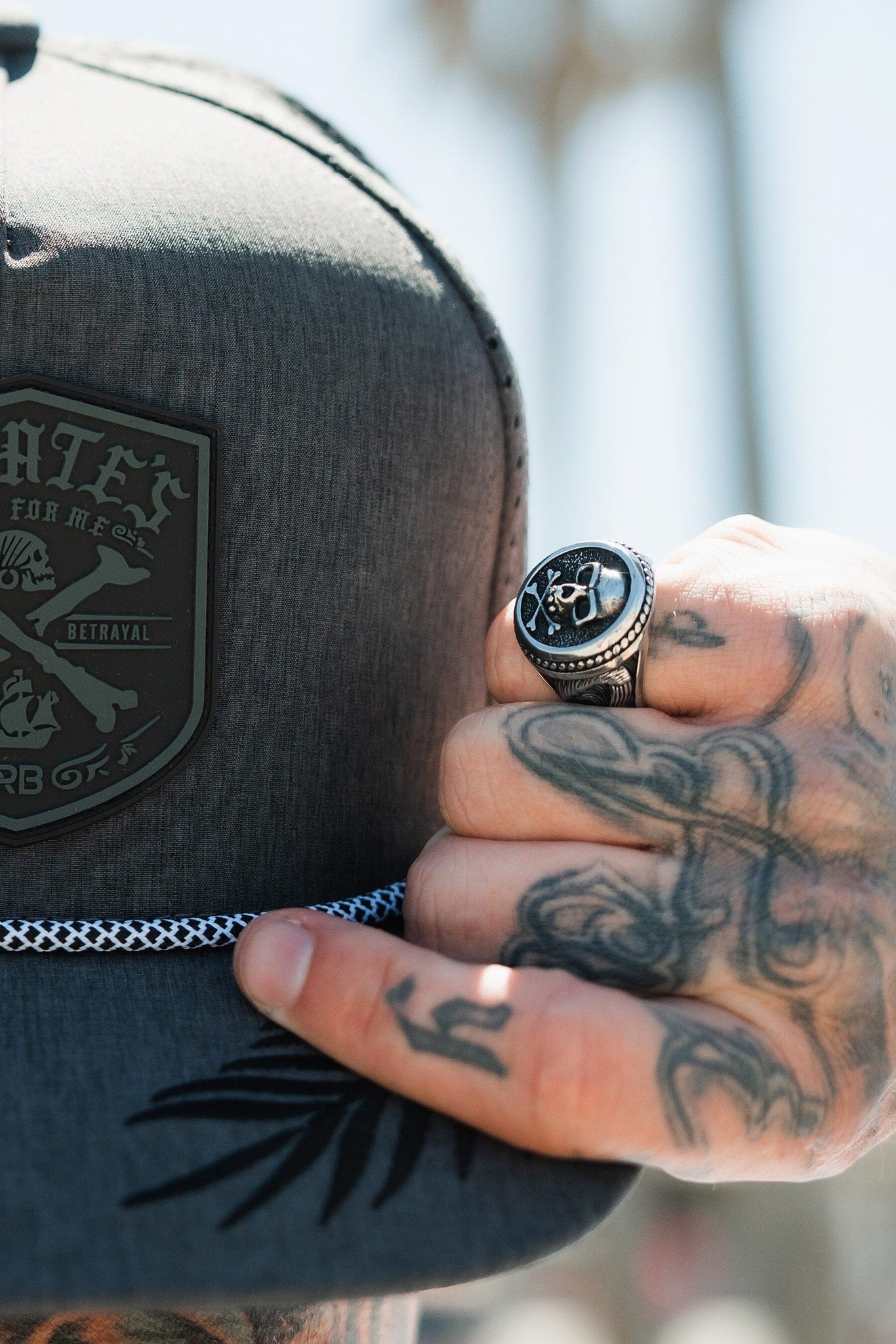 A tattooed hand adjusts a dark gray hat labeled with betrayal, reflecting the pirate lifestyle. Adorned on the hand is the handmade Pirates Life Stainless Steel Ring by Relentless Betrayal, with palm trees blurred in the background.