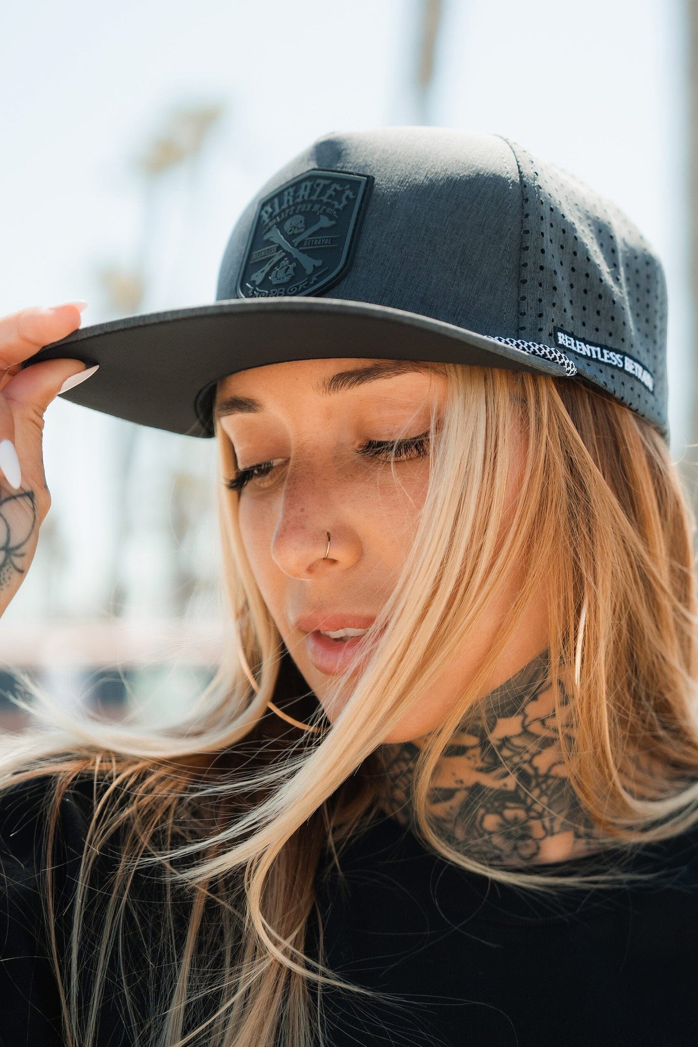 With tattoos and long hair, a person adjusts their Pirates Life Grey Snapback from Relentless Betrayal against a backdrop of blurred palm trees. They sport a nose piercing and the dark gray cap has a PVC rubber patch, embodying the pirates life vibe.