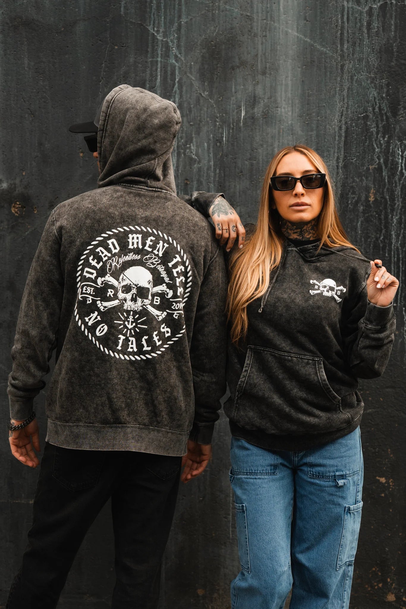 Two people in dark hoodies stand against a textured dark wall. The left showcases Relentless Betrayals Tell No Tales Premium Hoodie featuring a skull and Dead Men Tell No Tales on the back. The right faces forward, wearing sunglasses and blue jeans.
