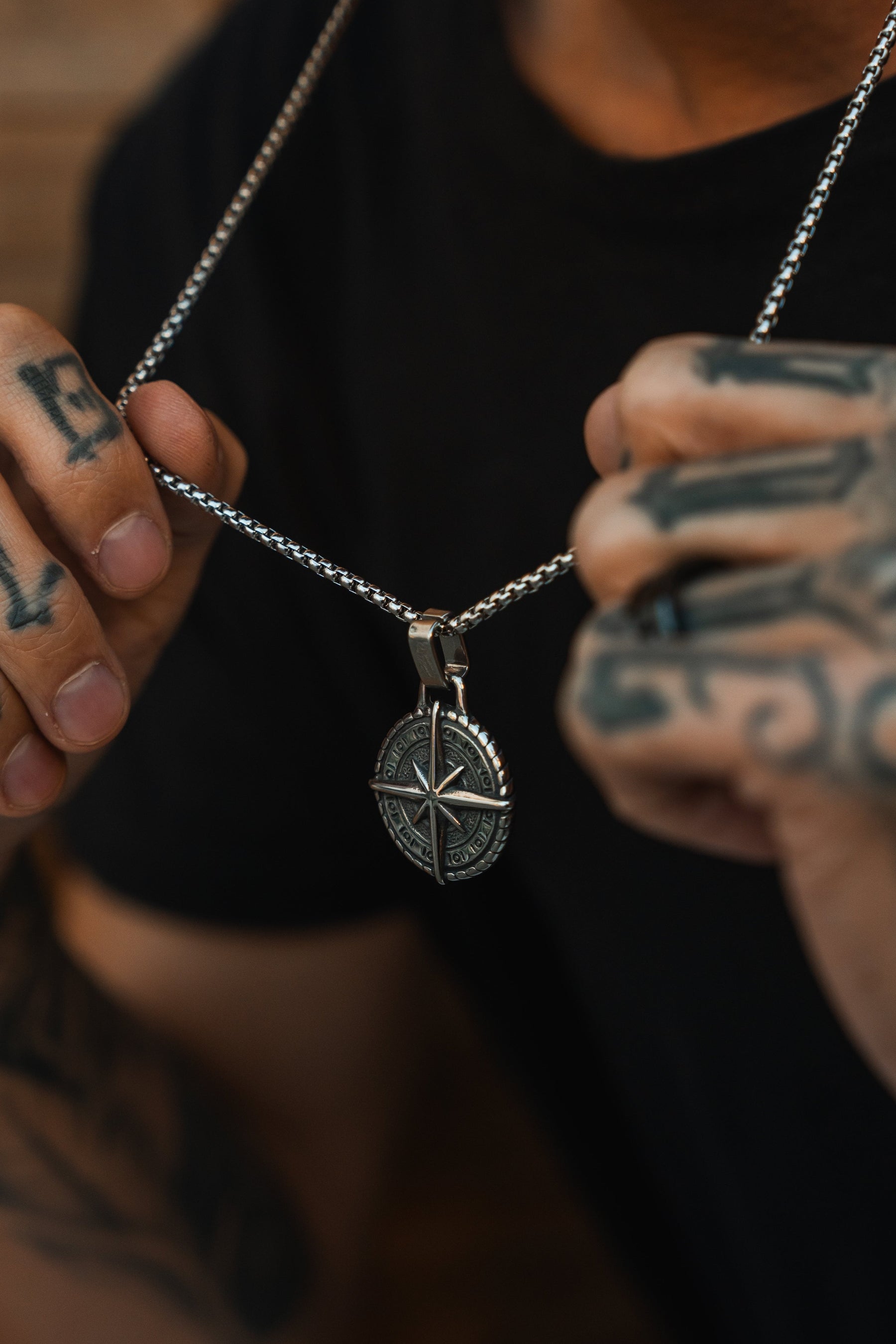 A person with tattooed hands showcases the dazzling Polaris Necklace by Relentless Betrayal. The intricate silver star-shaped pendant, adorned with sparkling gemstones, stands out against their black shirt, highlighting both the pendant and their vivid tattoos.