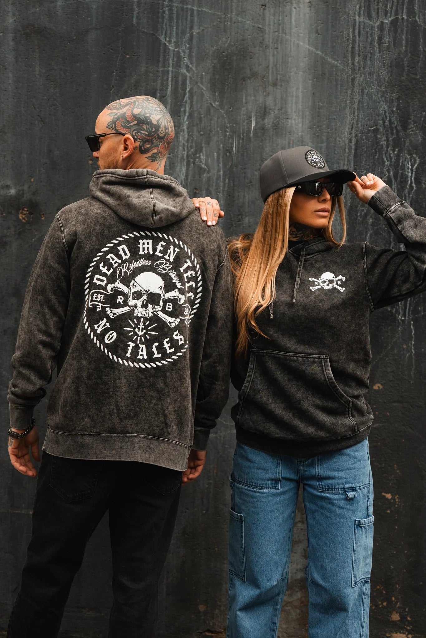 A man and woman lean against a dark, textured wall, wearing black Relentless Betrayal hoodies and sunglasses. The mans Tell No Tales Premium Hoodie showcases a Dead Men Tell No Tales skull design on the back, while the womans features a smaller skull on the front. Both wear caps with a mysterious coolness.