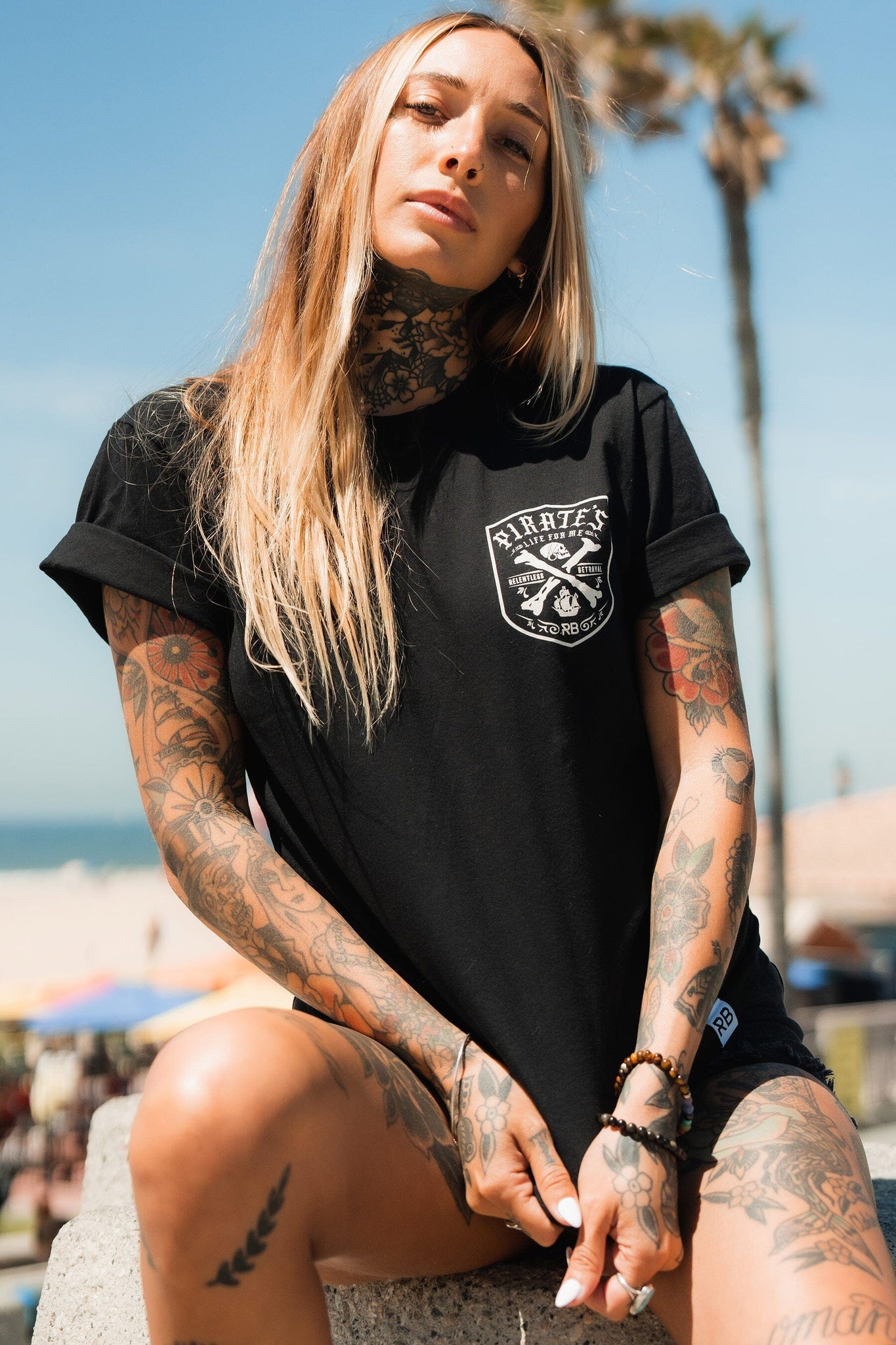 A tattooed woman with long blonde hair sits outdoors on a sunny day wearing a Pirates Life Black T-Shirt by Relentless Betrayal. Palm trees and beach scenes hint at a pirates adventure in the blurred background.