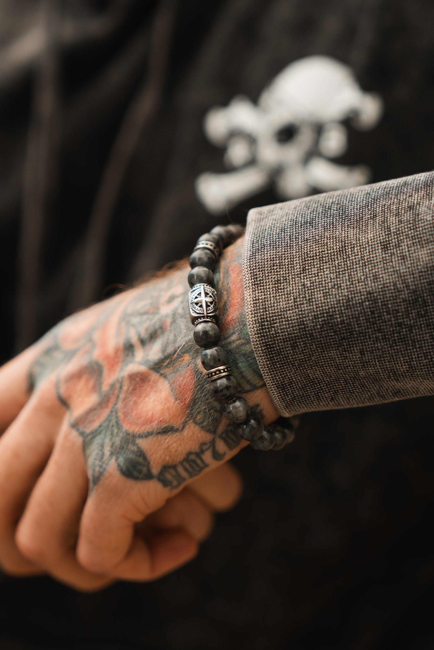 A tattooed hand with a Polaris Bead Bracelet by Relentless Betrayal graces the textured dark sleeve. A subtle skull and crossbones graphic is in the background, while a celestial Polaris Necklace sparkles nearby.