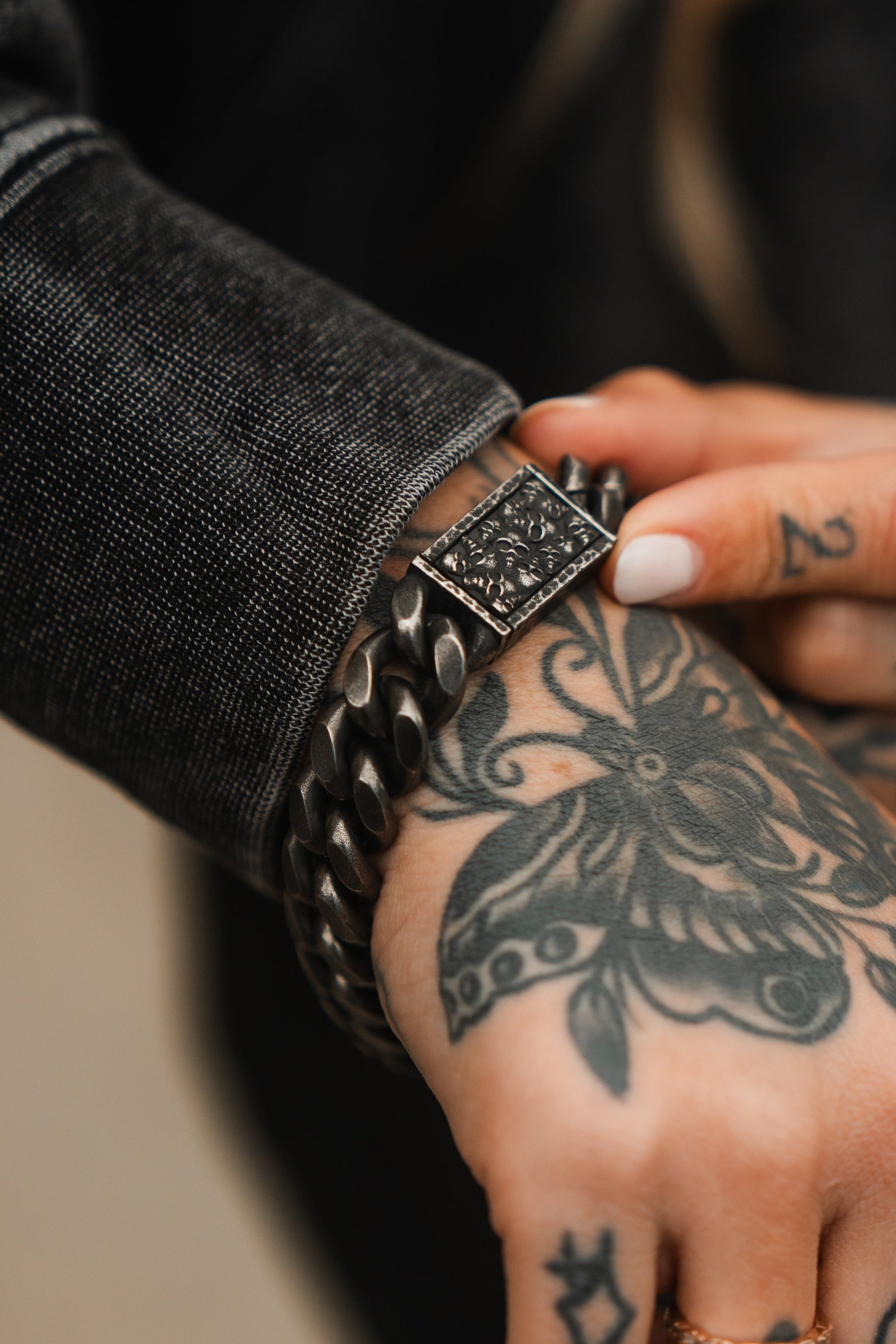 A person wearing a textured black jacket holds their wrist, adorned with the Tell No Tales Bracelet by Relentless Betrayal, featuring an ornate clasp. Their hand and wrist display floral and abstract tattoos, while short white-painted nails complete their edgy style.