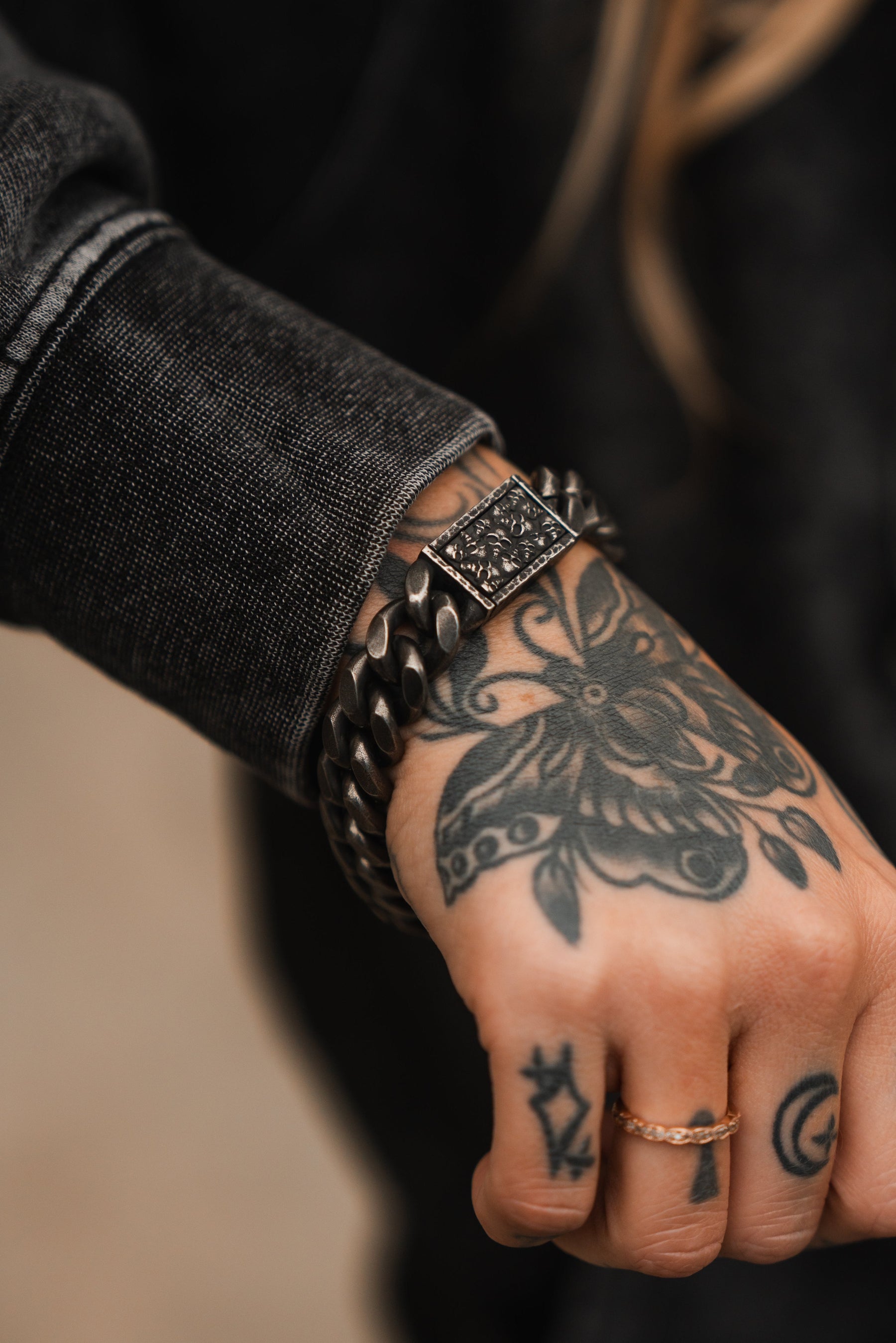 A hand adorned with floral tattoos showcases a Tell No Tales Bracelet by Relentless Betrayal. Complementing the distinct look, this person wears a classic black denim jacket.