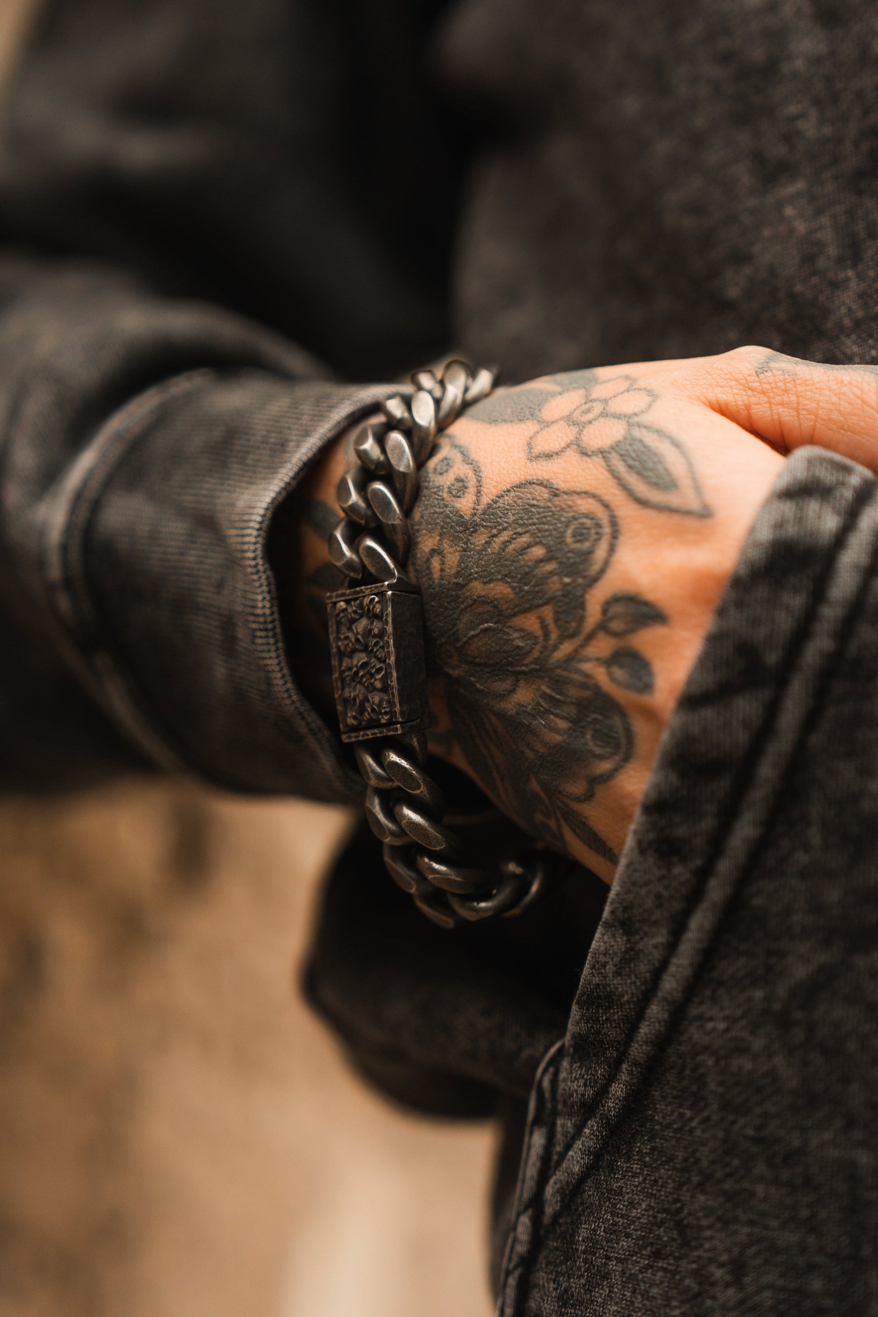 A hand with floral tattoos rests in a gray denim pocket, adorned by the Tell No Tales Bracelet from Relentless Betrayal. Hand-forged stainless steel with an ornate rectangular clasp and lifetime warranty, it adds a bold touch to the casual outfit against a softly blurred background.