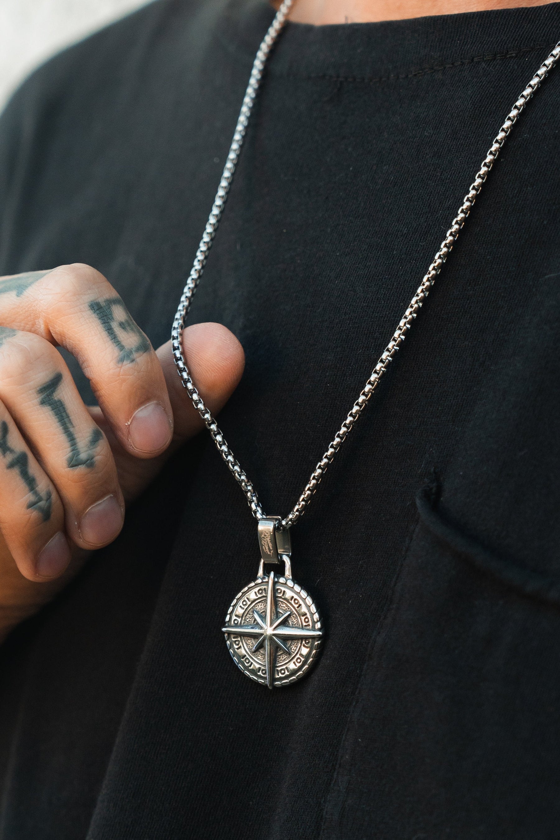A tattooed person in a black shirt holds the Relentless Betrayal Polaris Necklace, featuring a silver compass pendant with star-shaped circular details, elegantly hanging on its simple chain.