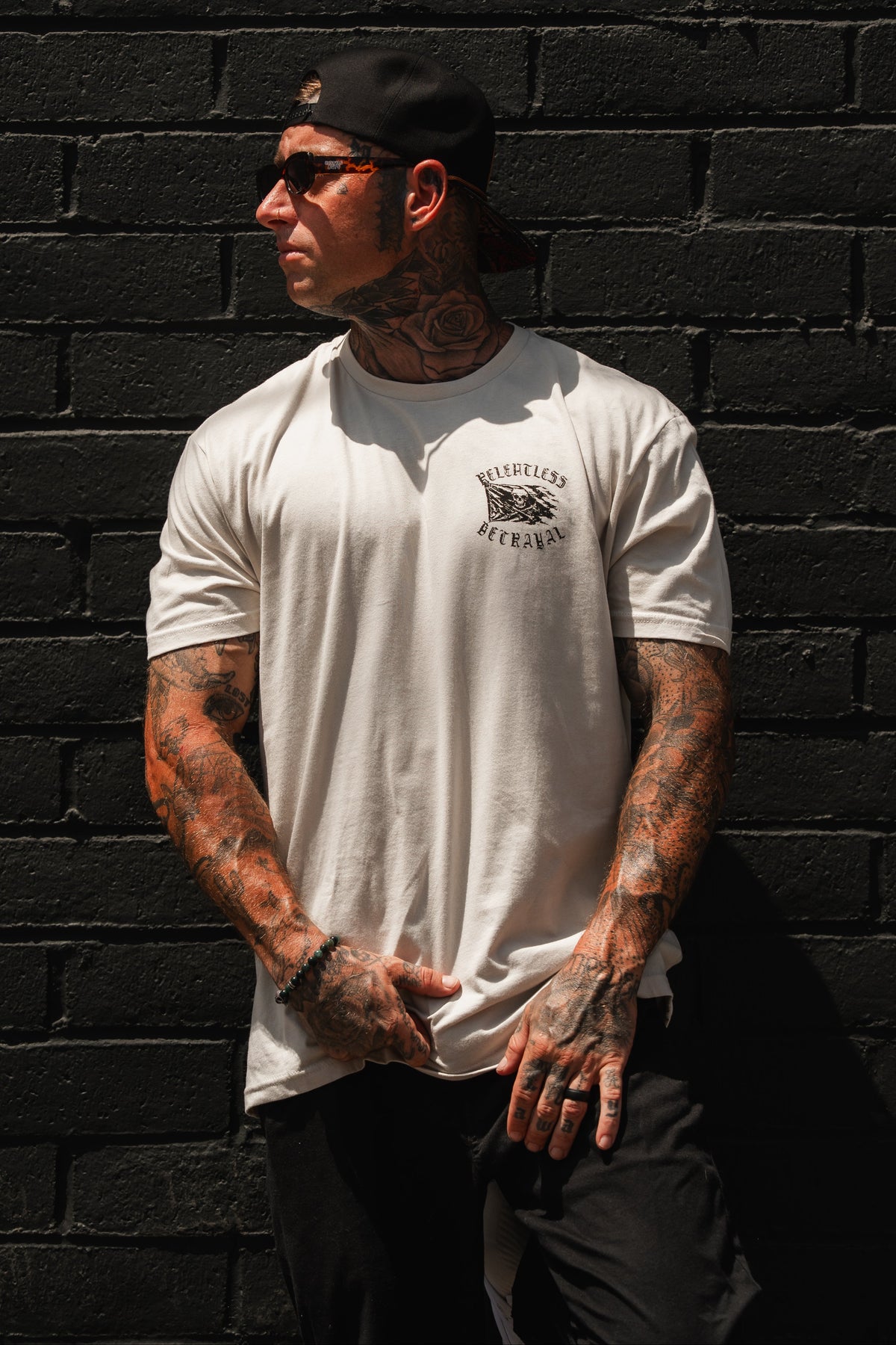 A tattooed man wearing the Raise the Jolly Roger Bone T-Shirt from Relentless Betrayal, along with a black cap and sunglasses, stands against a black brick wall, looking to the side.