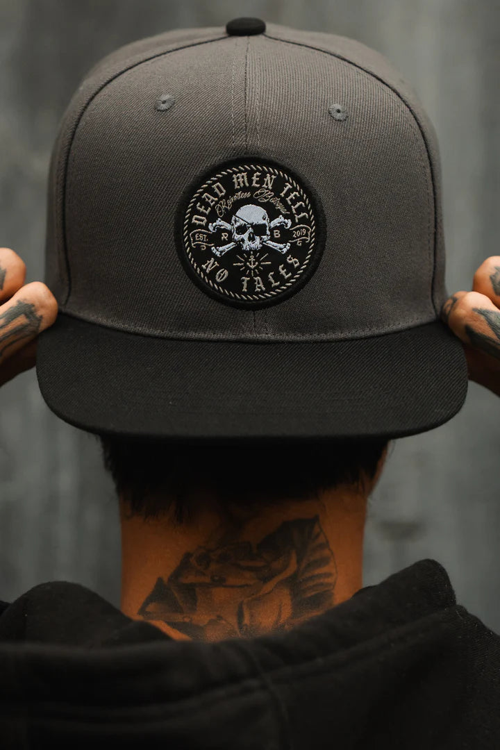 A tattooed individual wears a Relentless Betrayals Dead Men Tell No Tales snapback, featuring a circular skull and crossbones patch. Made from a soft polyester cotton blend, the gray handmade cap is stylishly set against a gray background.