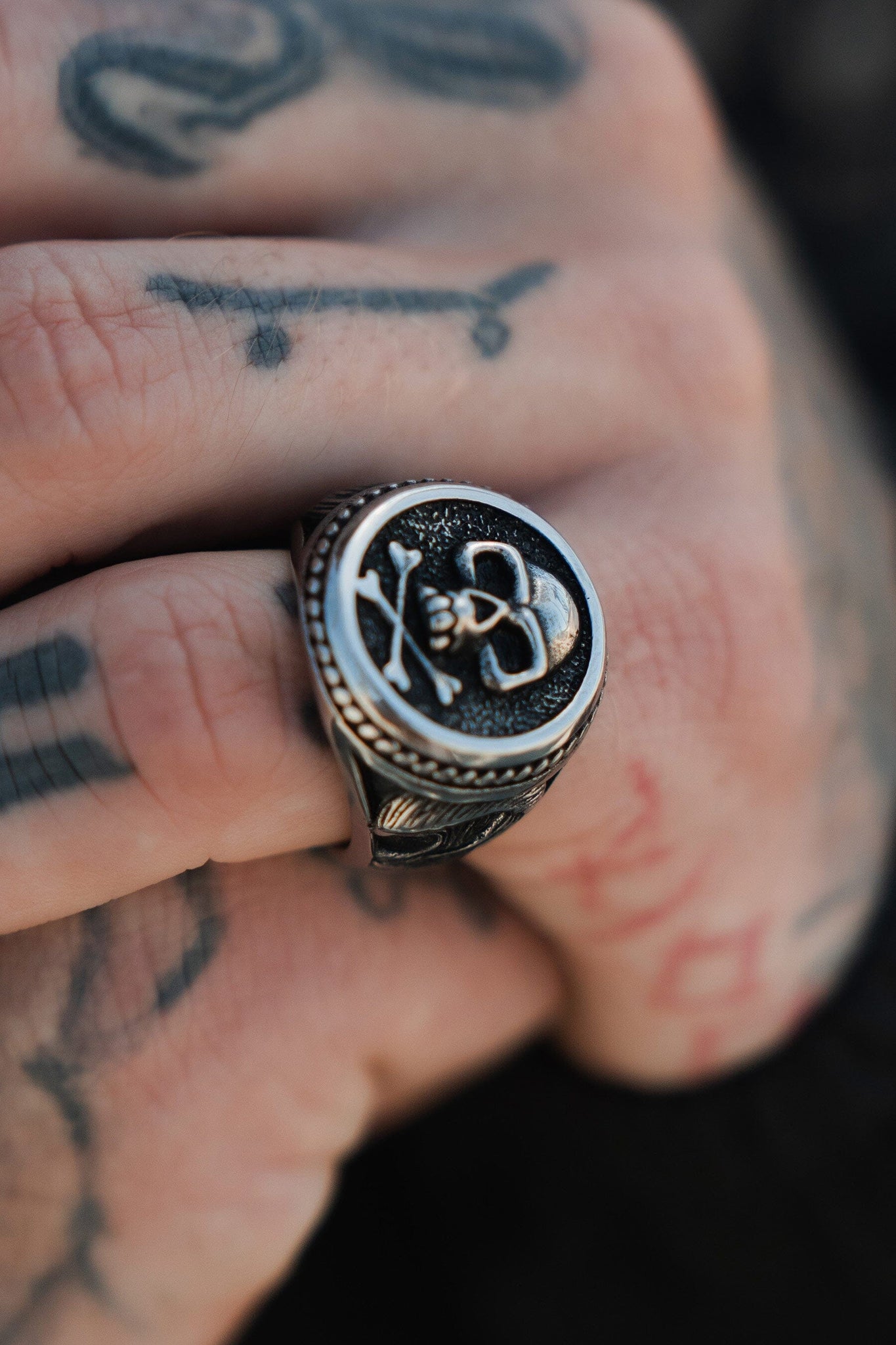 A tattooed hand on a textured, dark surface wears Relentless Betrayals Pirates Life Stainless Steel Ring. The handcrafted skull design adorns the middle finger, evoking the pirate lifestyle with its intricate details and craftsmanship.