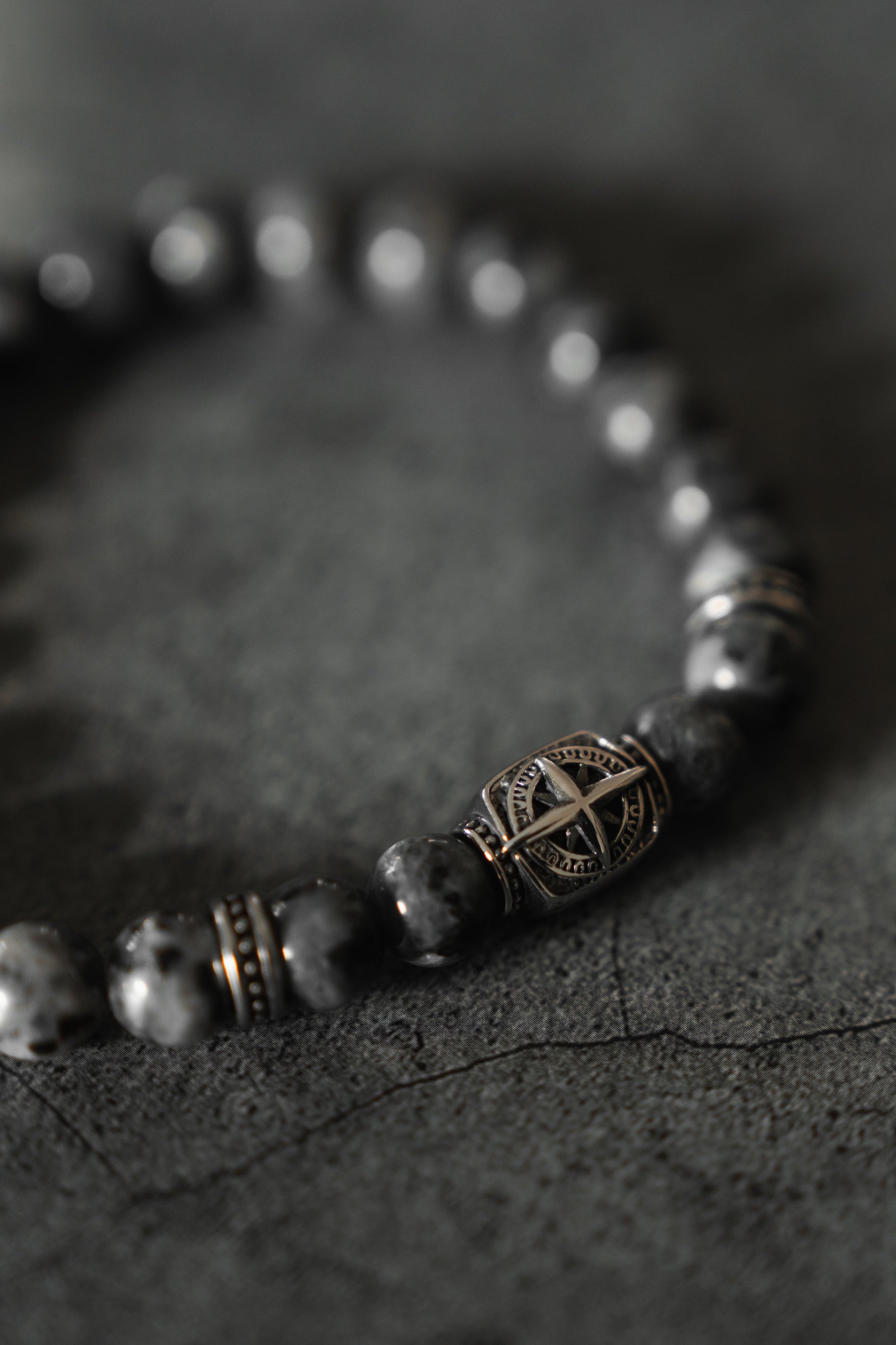 A close-up shot of the Polaris Bead Bracelet by Relentless Betrayal showcases its dark, marbled beads on a textured surface. The centerpiece features a star-shaped pattern with a metallic finish, providing an intricate contrast to the bracelets smooth, glossy beads.