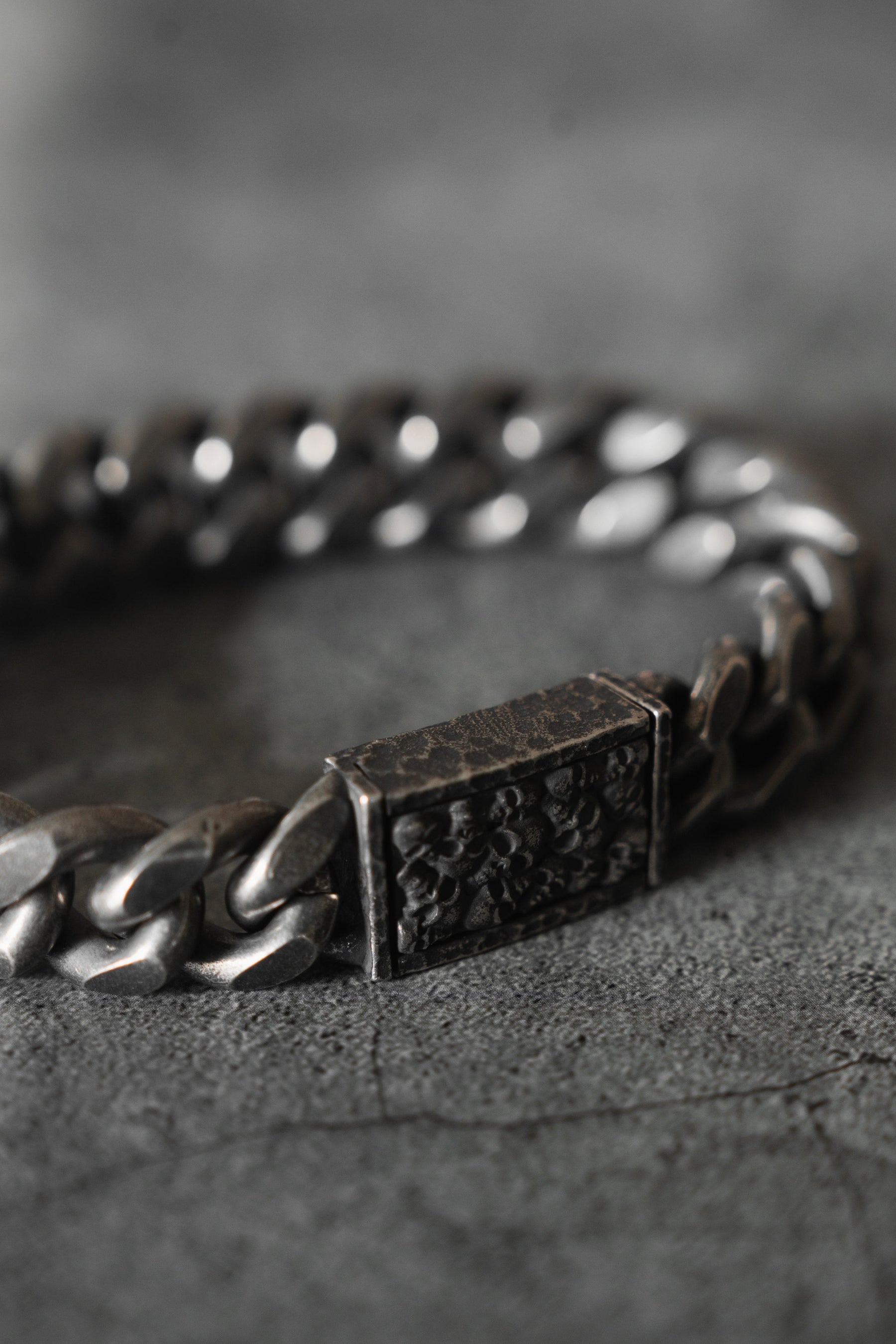 The Tell No Tales Bracelet by Relentless Betrayal is showcased up close on a textured dark gray surface. This hand-forged silver and stainless steel bracelet features a rectangular clasp with intricate carvings, emphasizing its detailed design and lifetime warranty-backed durability.