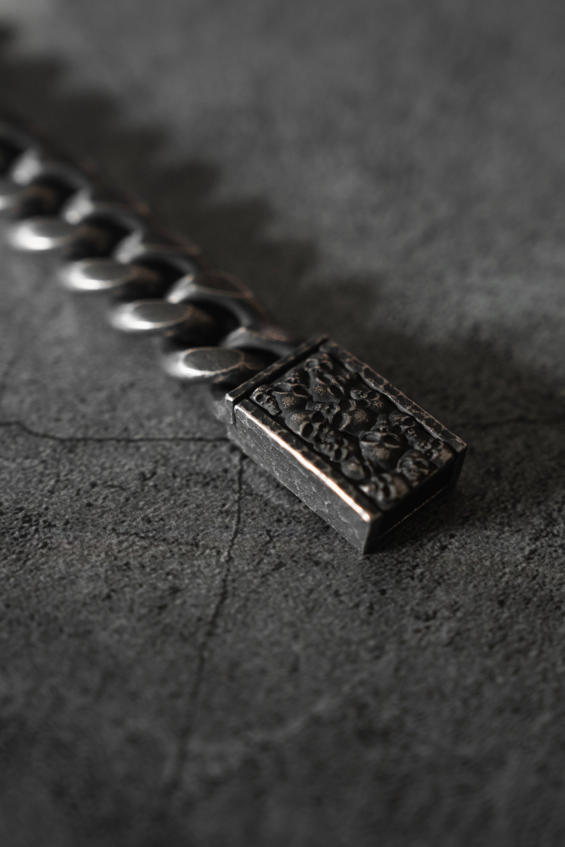 A Tell No Tales Bracelet by Relentless Betrayal, featuring a hand-forged silver chain and intricate rectangular clasp, lies on a dark, cracked surface. The close-up captures the bracelets craftsmanship and timeless elegance.