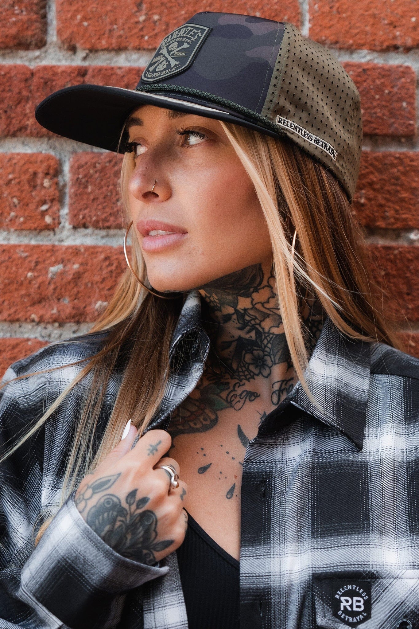 Against a brick wall, someone embodies adventure wearing a water-resistant Pirates Life Camo Snapback by Relentless Betrayal and a plaid shirt. Tattoos adorn their neck and hand, hoop earrings dangle from their ears, and a nose ring adds edge as they gaze sideways.