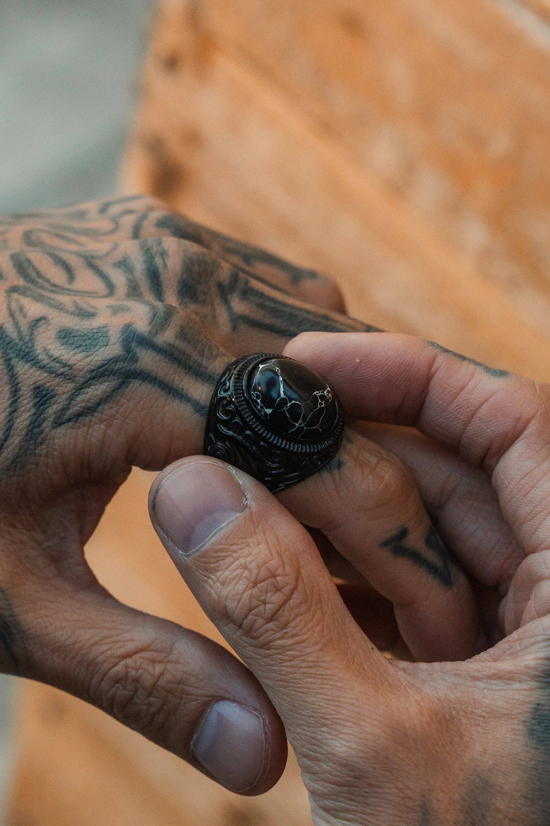 A person wears The Dark Storm Ring by Relentless Betrayal, its dark design contrasting with intricate tattoos. The stainless steel band subtly gleams against the blurred backdrop, enhancing its mystical aura amidst a warm wooden texture.