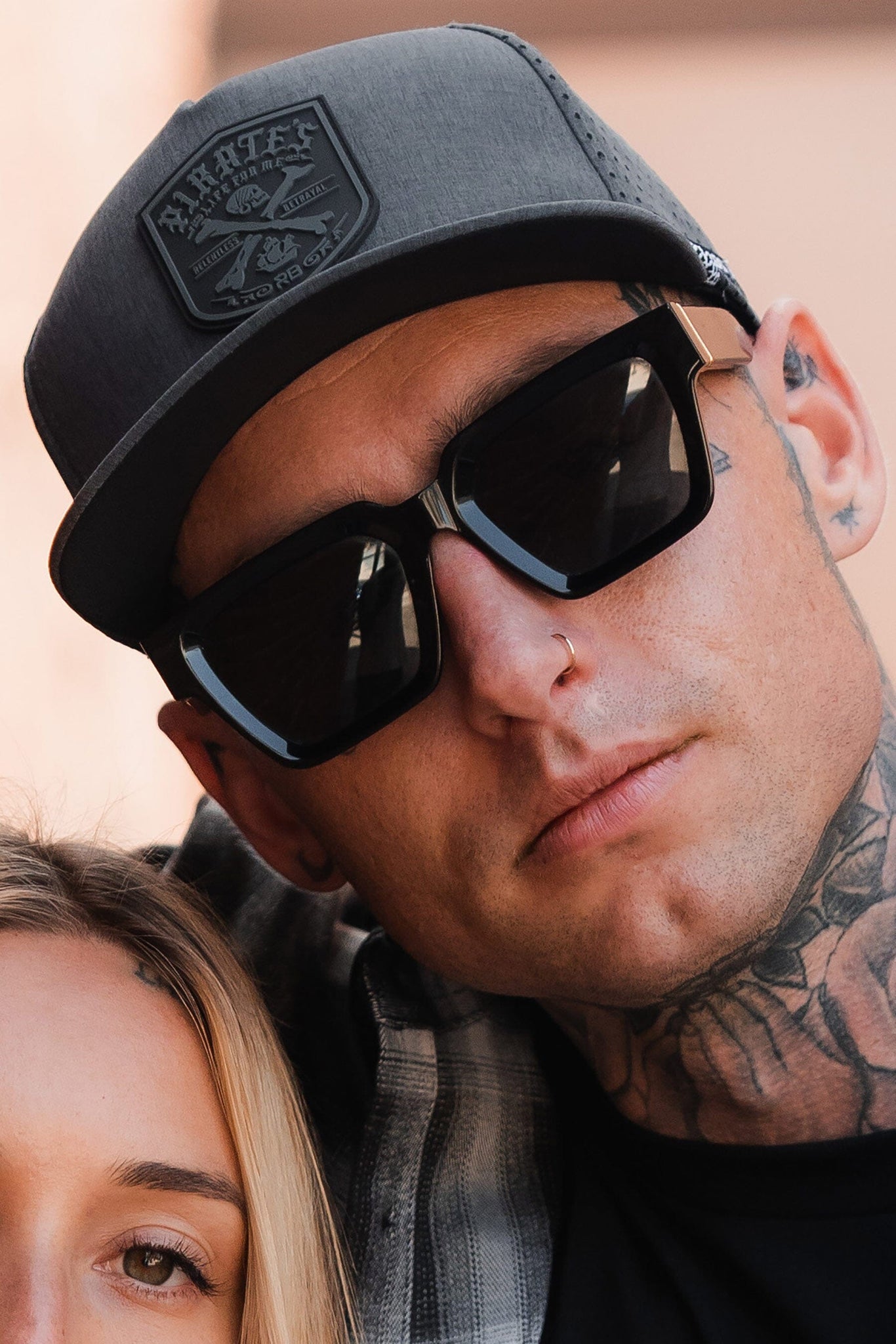 A man in a water-resistant Pirates Life Grey Snapback by Relentless Betrayal, large sunglasses, and a plaid shirt gazes forward. He has tattoos and a nose piercing. Beside him, part of another face is visible, enhancing the mysterious allure of this modern-day snapshot.