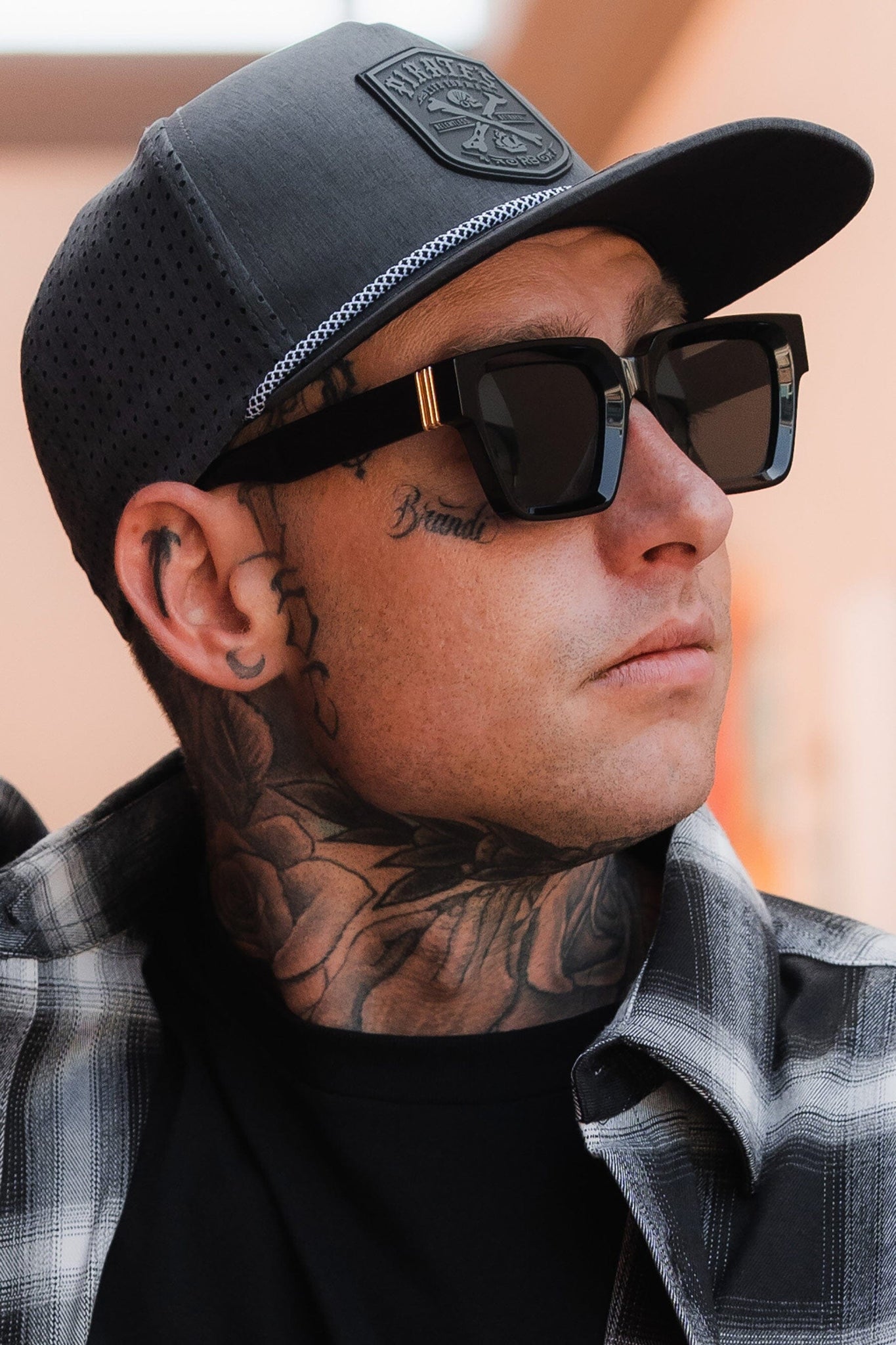 A person in a Pirates Life Grey Snapback by Relentless Betrayal gazes sideways. They have tattoos on their neck and face, exuding rugged charm in a plaid shirt over a black top.