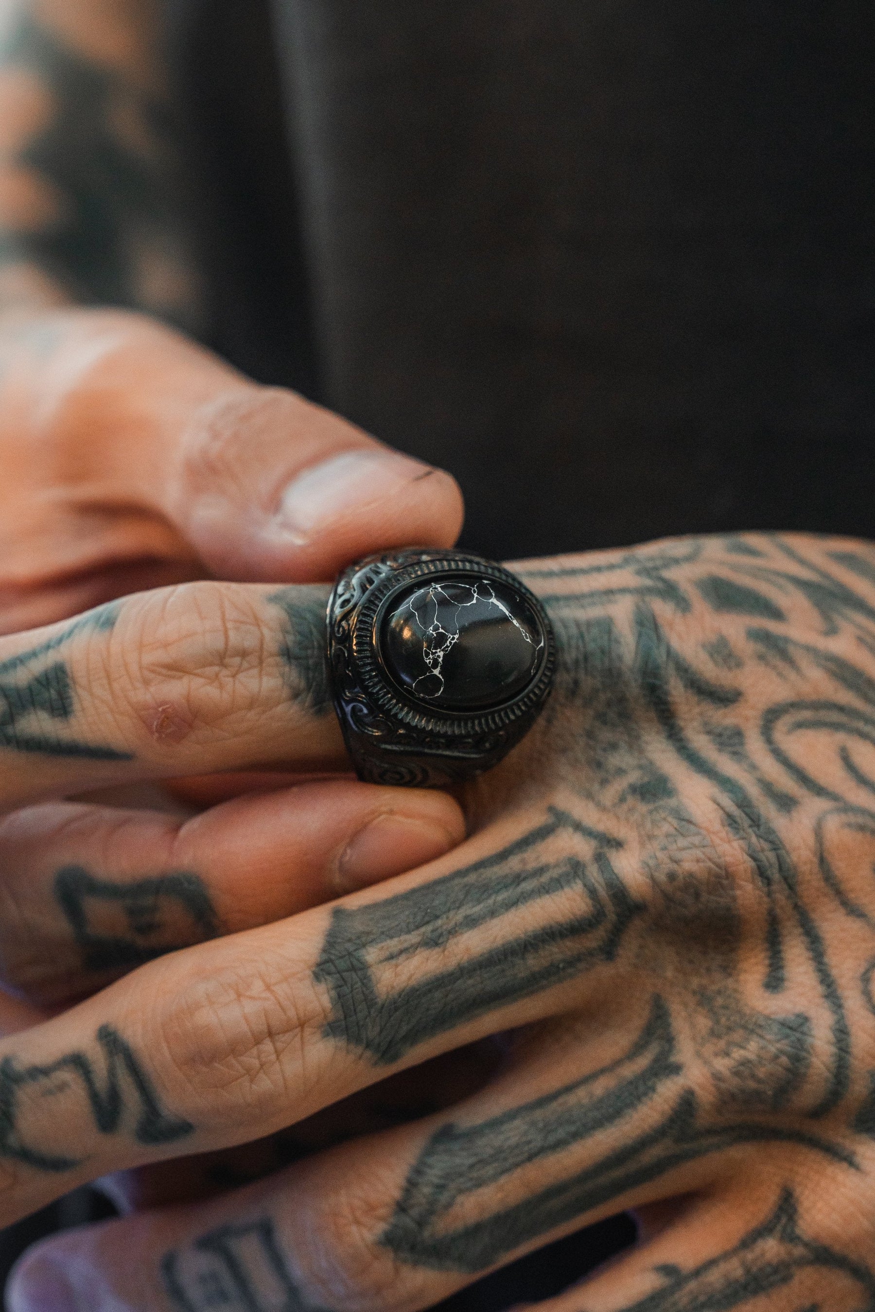 A tattooed hand showcases The Dark Storm Ring by Relentless Betrayal, an ornate stainless steel piece with a dark polished stone engraved with a constellation. Other hand tattoos are visible in the background.