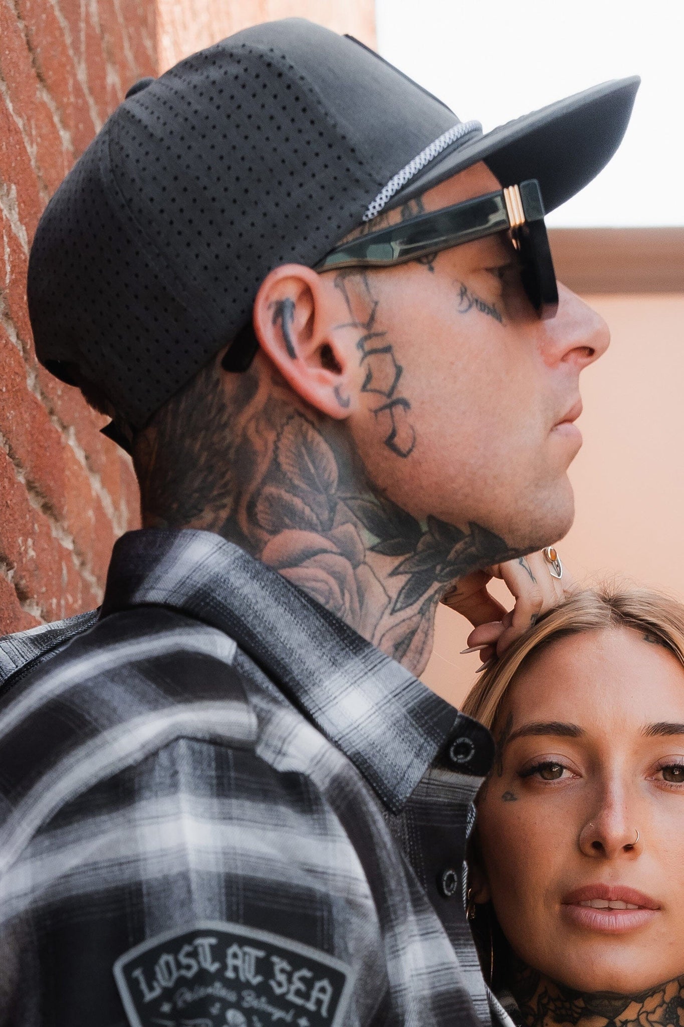 A person wearing the Pirates Life Grey Snapback by Relentless Betrayal, sunglasses, and a plaid shirt stands against a brick wall, showcasing neck and face tattoos. Another person leans on their shoulder, gazing at the camera—capturing an effortless pirates life moment.