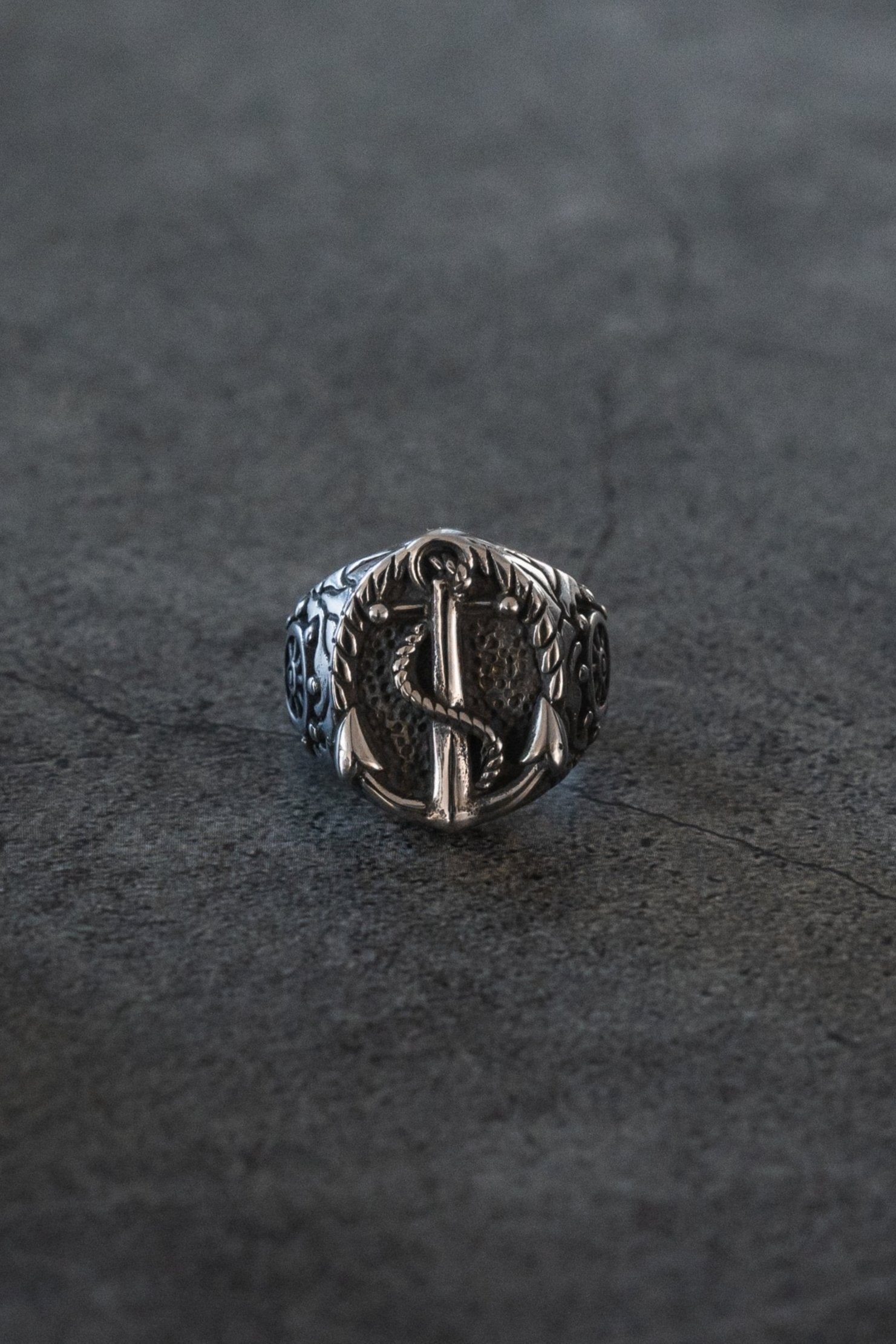 The Voyager Ring by Relentless Betrayal is made from stainless steel, showcasing an anchor with rope and laurel leaves on a dark textured surface. The band features intricate swirling patterns, perfect for adventure and exploration jewelry enthusiasts.