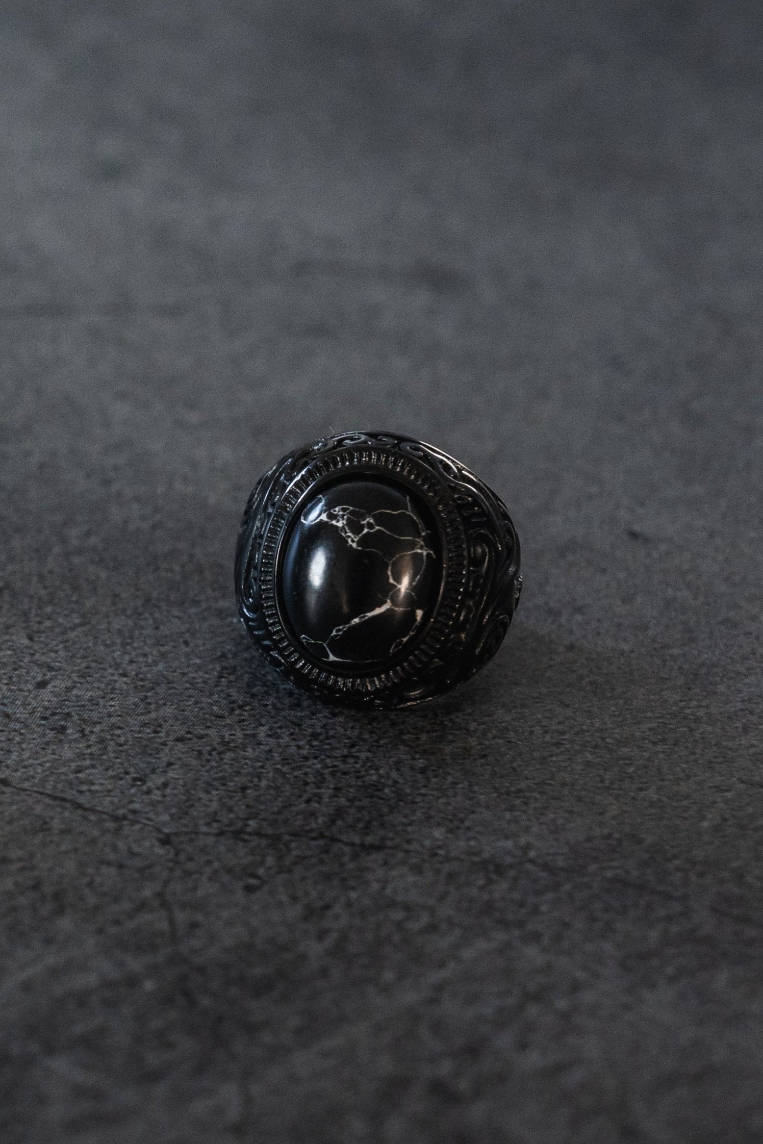 The Dark Storm Ring by Relentless Betrayal showcases an oval marbled stone on a textured gray surface, with a black stainless steel band featuring intricate designs that enhance the stones glossy finish.