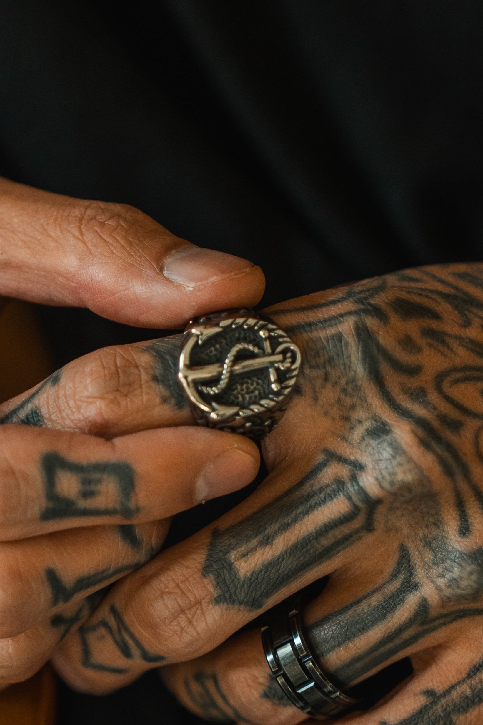 Tattooed hands adjust a detailed silver Voyager Ring with a snake design by Relentless Betrayal. The fingers, adorned with a black band ring, are intricately tattooed, embodying the adventurous spirit of exploration jewelry.