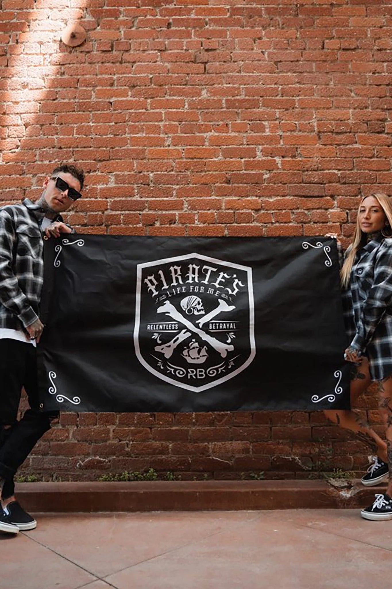 Two people in black and white plaid shirts stand against a brick wall, holding the Relentless Betrayals durable polyester Pirates Life Flag, featuring a skull and bones emblem.
