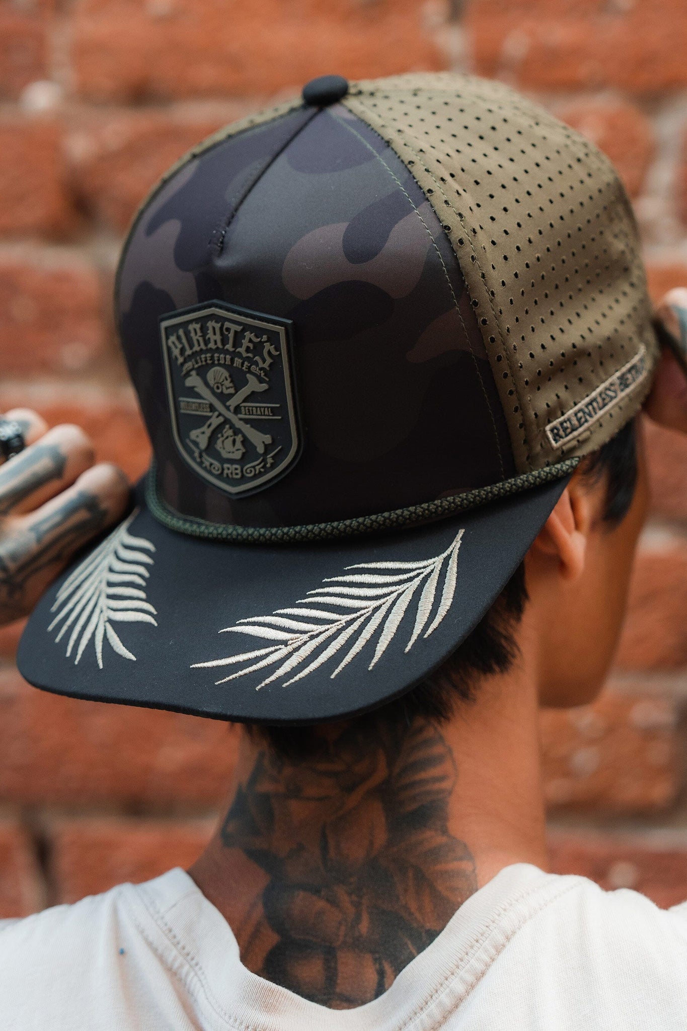 With tattoos adorning their neck and hands, they sport the Pirates Life Camo Snapback by Relentless Betrayal. This water-resistant hat features a pirate patch, embroidered palm leaves, and RELENTLESS HONOR on the side against a brick wall backdrop, embodying an adventurous spirit.