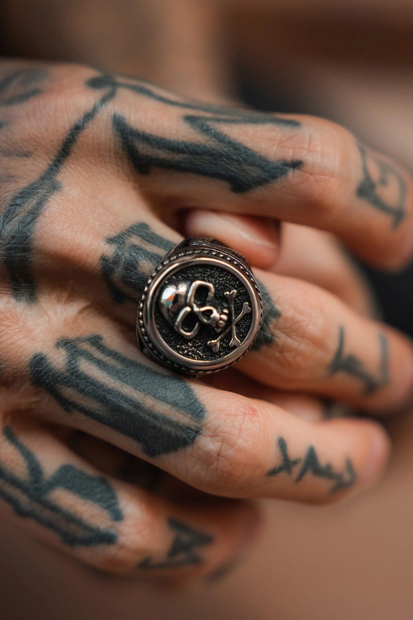 A tattooed hand displays a Relentless Betrayals Pirates Life Stainless Steel Ring, featuring an embossed floral design that exudes a pirate lifestyle. Abstract tattoos enhance the contrast against the rings metallic sheen.