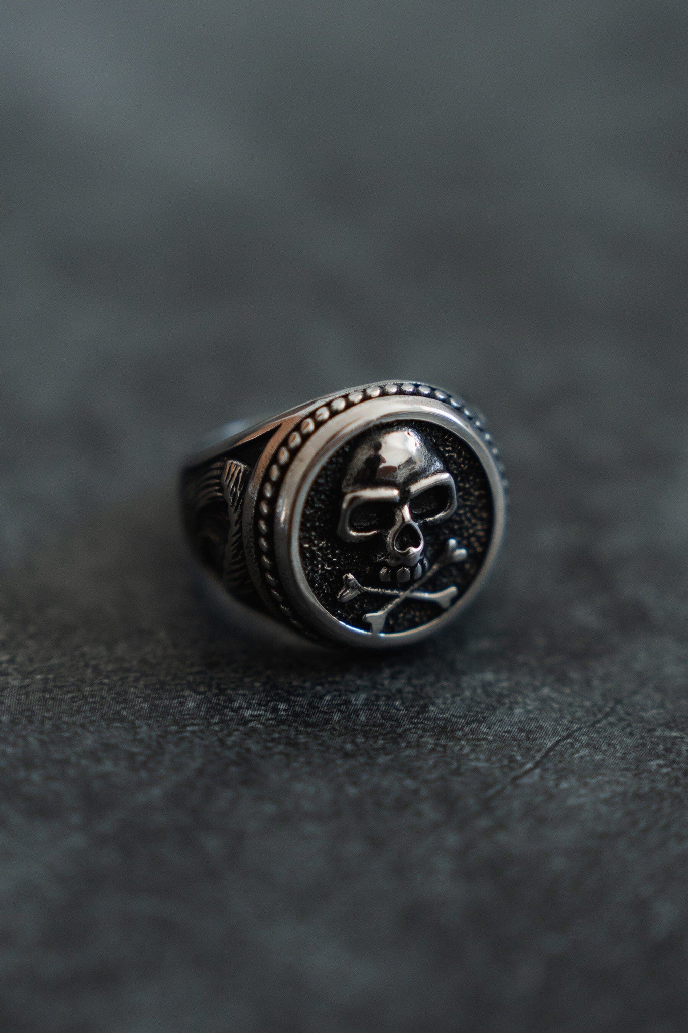 The Pirates Life Stainless Steel Ring by Relentless Betrayal features a raised skull and crossbones on a round face with detailed engravings, embodying the pirate lifestyle. Perfect for those living by Pirates Life For Me, this ring sits strikingly on a gray surface.