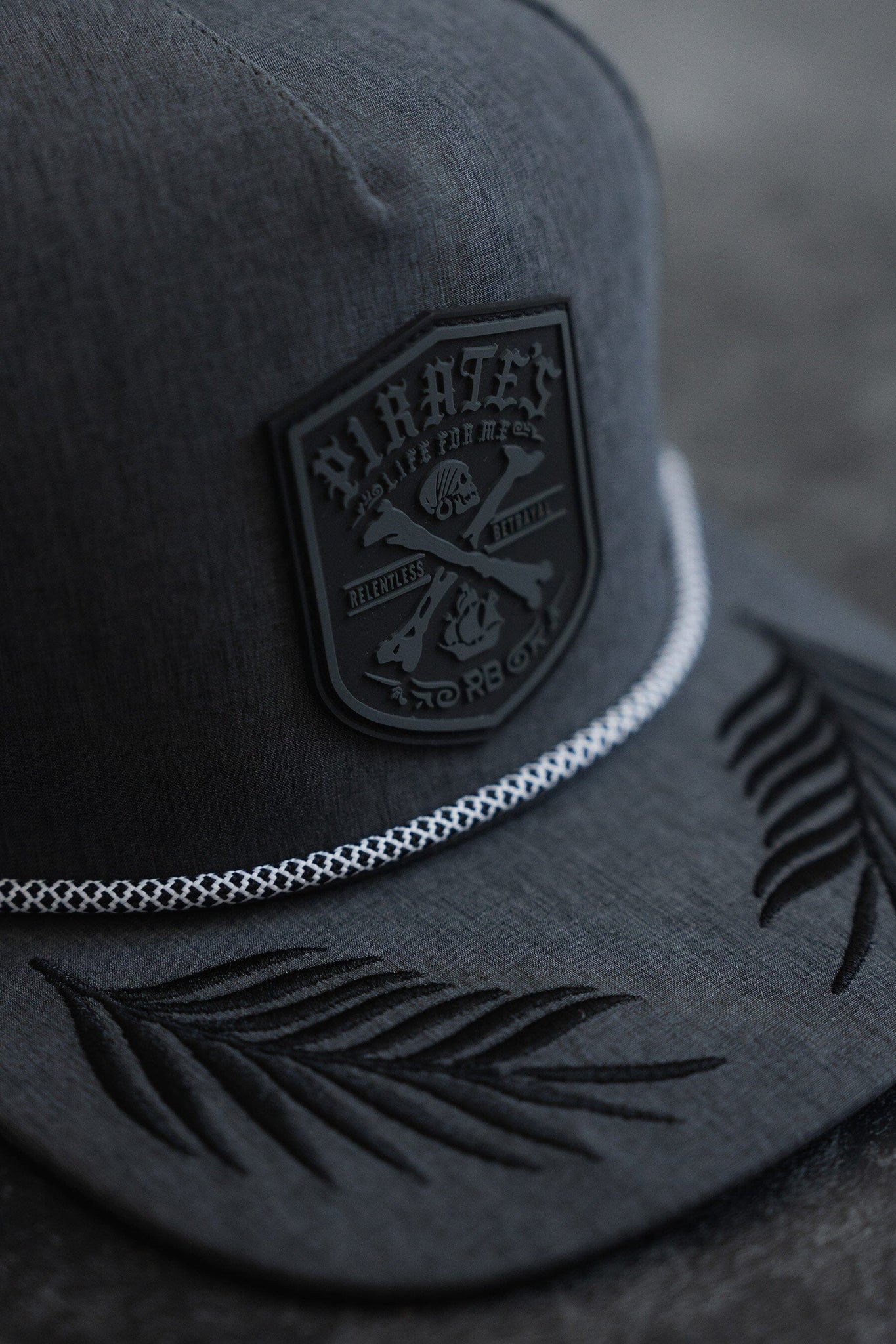 The Pirates Life Grey Snapback by Relentless Betrayal features a dark gray, water-resistant design with a PVC rubber patch of graphics and text. It includes a decorative white rope above the brim, with embroidered leaf patterns on the brim against its dark, textured surface.