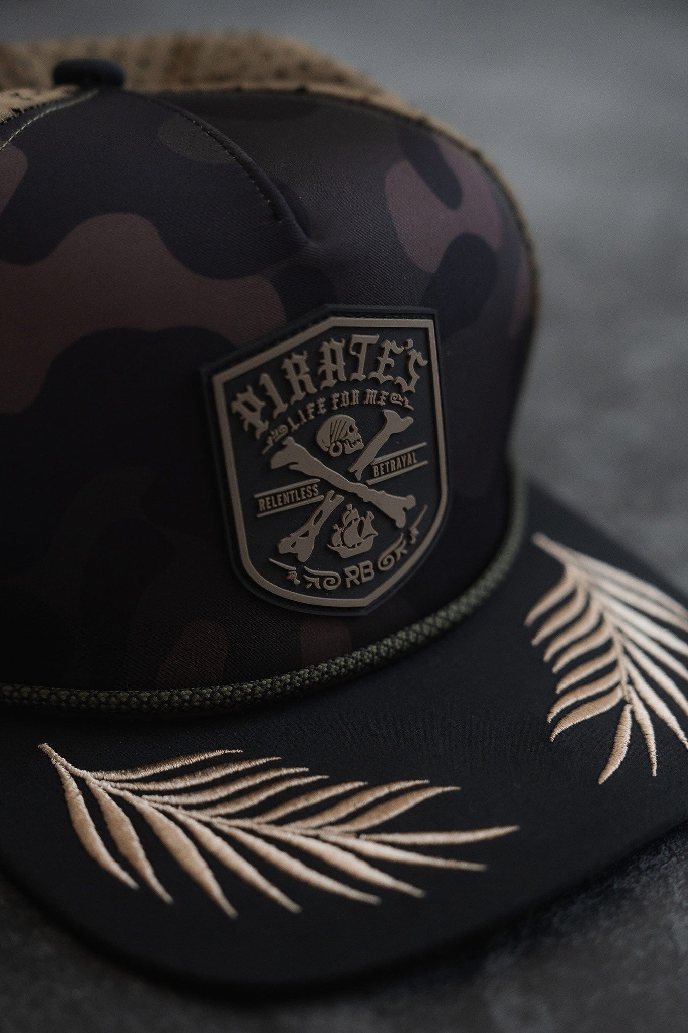 The Pirates Life Camo Snapback by Relentless Betrayal features a patch with Pirates, Life for Me, Relentless, Betrayal and skull and crossed bones flanked by pistols. This water-resistant hat is perfect for adventure seekers with beige embroidered palm leaves on the brim and a green braided trim.