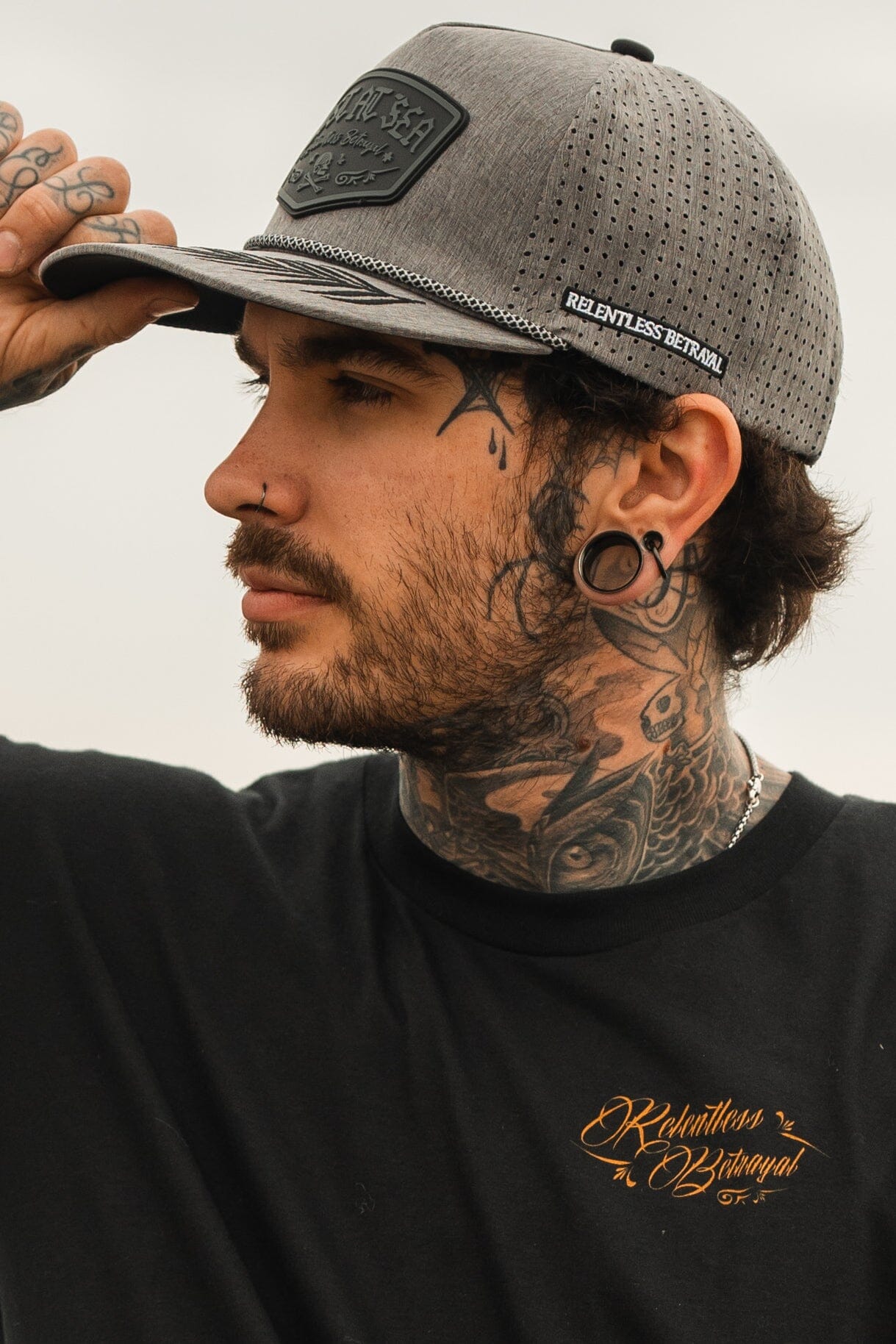 A man with tattoos is wearing a Lost At Sea Gunmetal PVC Snapback by Relentless Betrayal, complementing his large gauged earrings, nose ring, and a black shirt with orange text as he gazes to the right.