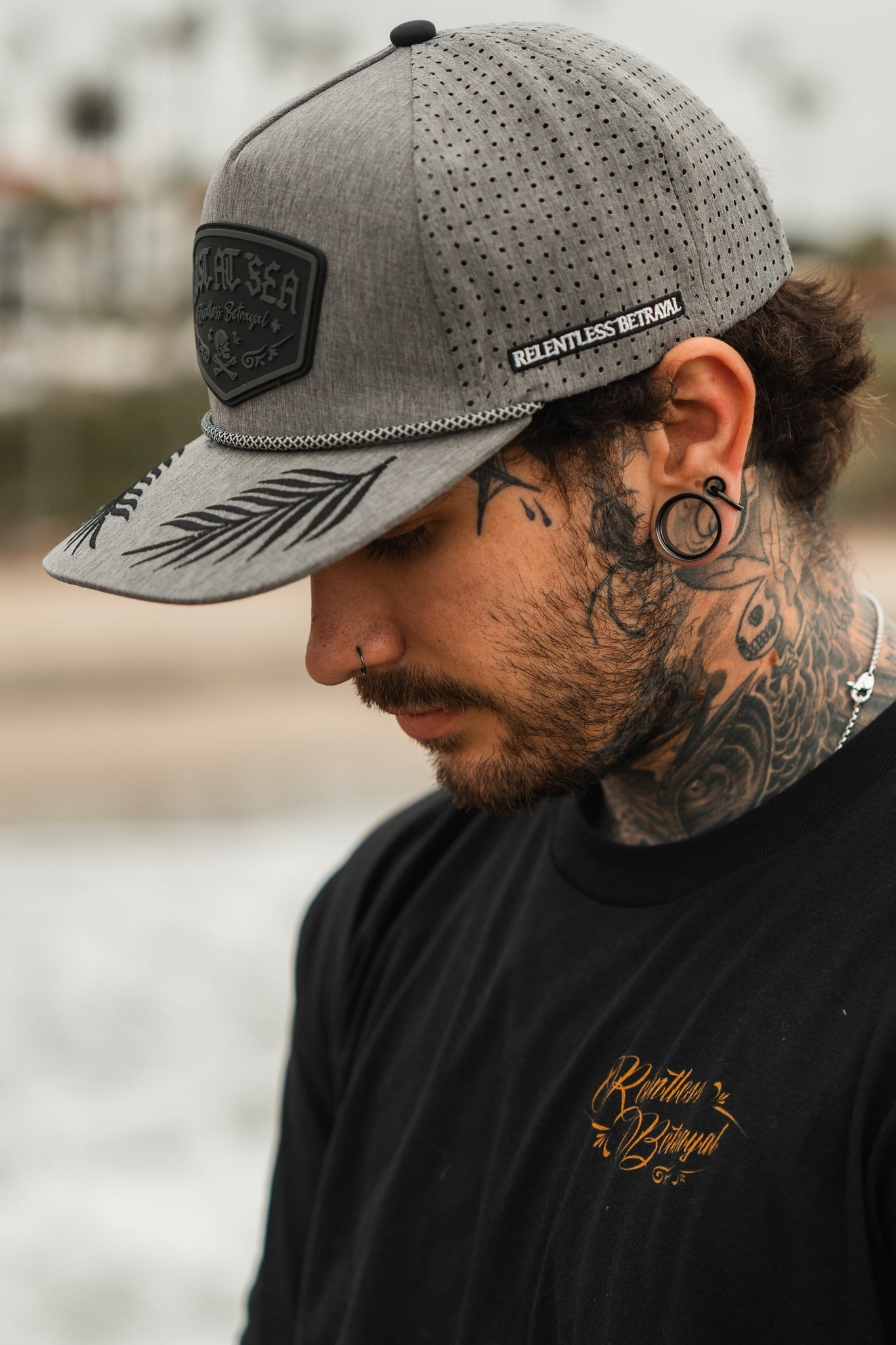 A man with neck tattoos and ear gauges wears the Lost At Sea Gunmetal PVC Snapback by Relentless Betrayal. He has a nose piercing and dons a black shirt with gold writing, enhancing his style as he stands outdoors against a blurred backdrop.