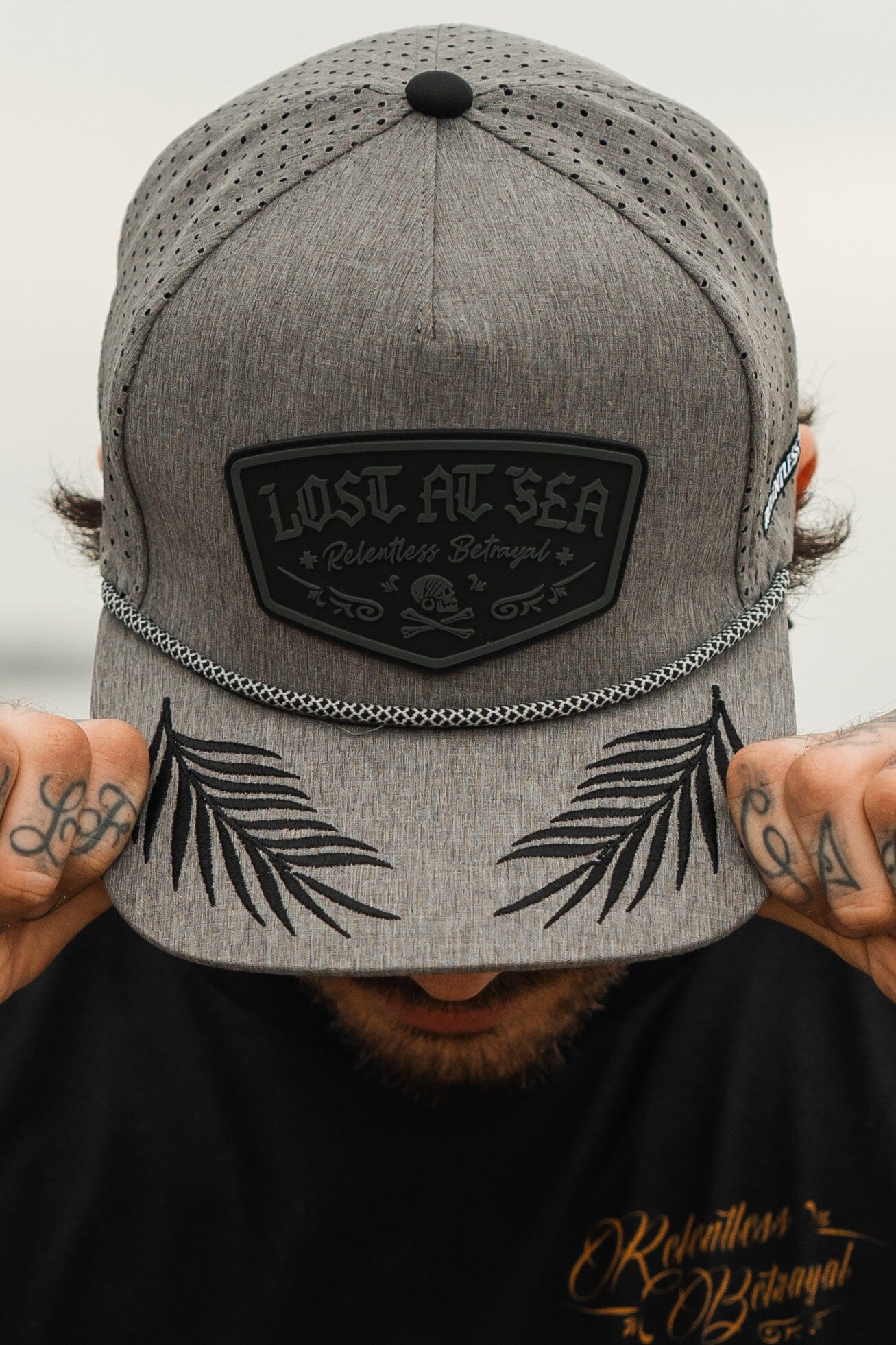 A person wears the Lost At Sea Gunmetal PVC Snapback by Relentless Betrayal, featuring a skull design and black leaf patterns on the brim. They adjust the water-resistant hat with both hands, partially covering their face.