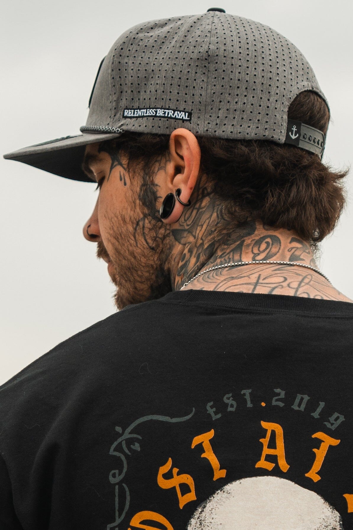 Facing away, an individual sports the Lost At Sea Gunmetal PVC Snapback from Relentless Betrayal and a black T-shirt with a partial design. They have ear gauges and visible neck tattoos, with RELENTLESS BETRAYAL displayed on the caps back.