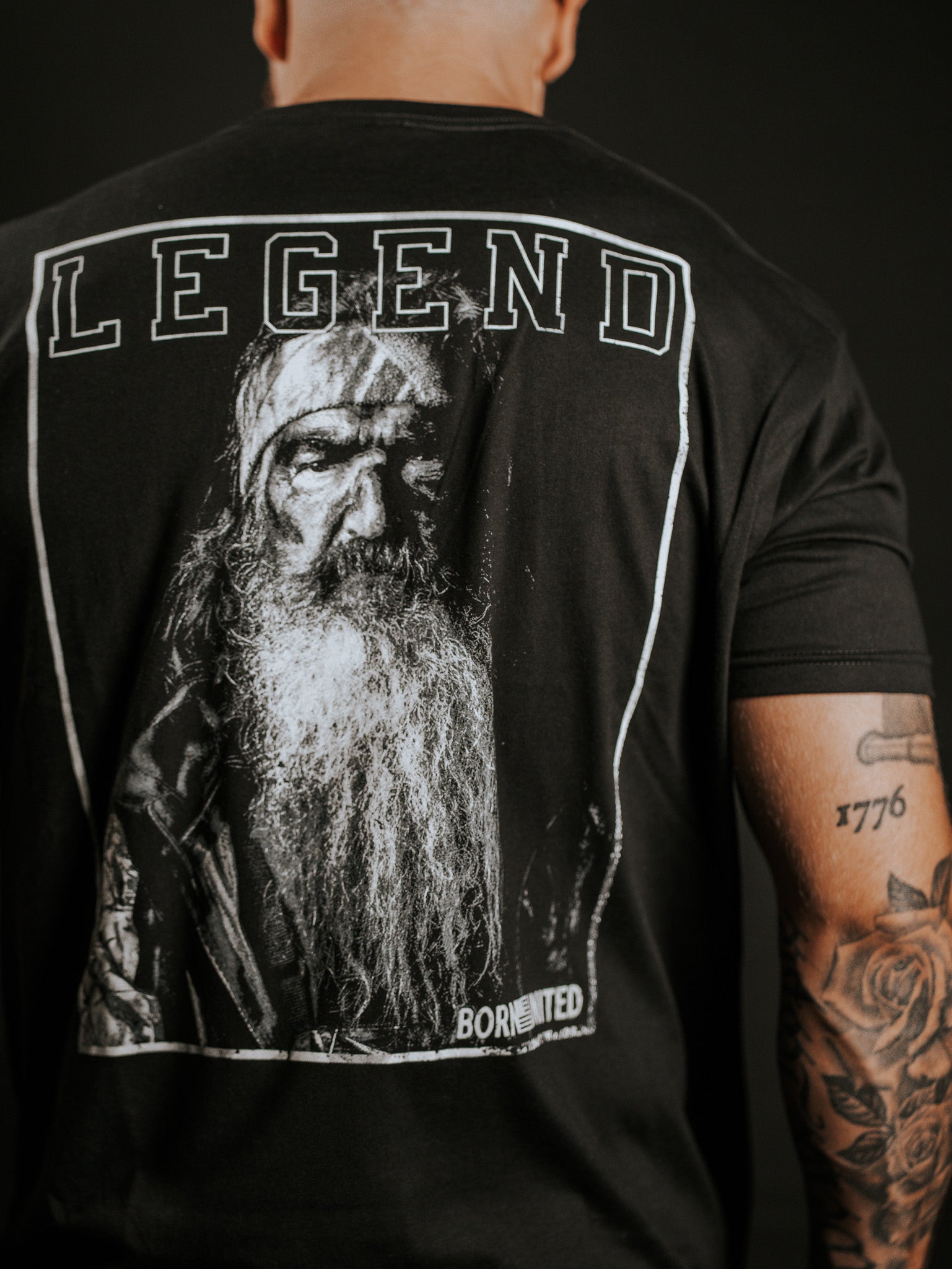 A person with tattooed arms wears the Legend Dynasty T-shirt by Born United Private Label, showcasing a large grayscale portrait of an older man with a long beard and LEGEND printed above the image.