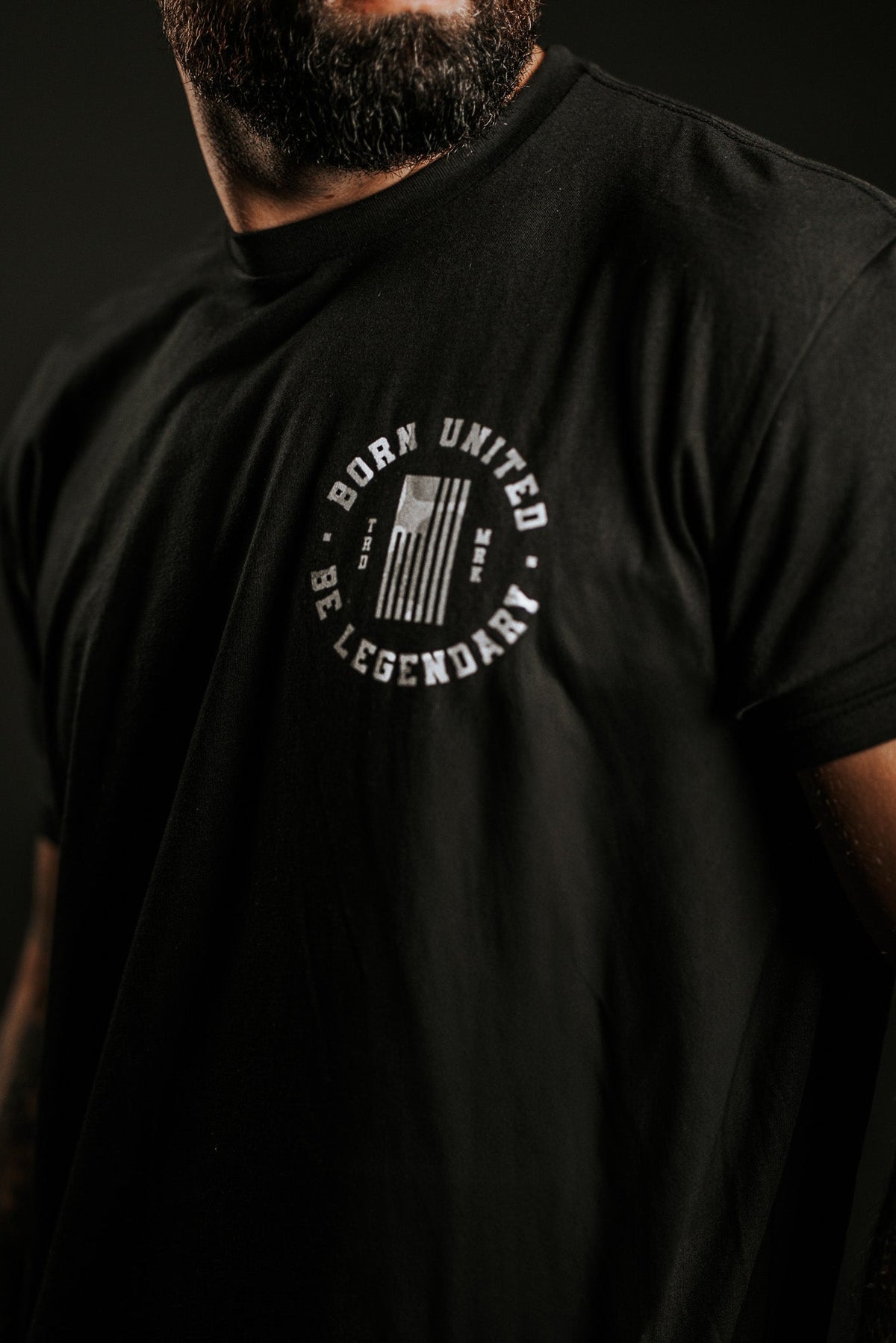 A bearded man wears a black Legend Dynasty t-shirt by Born United Private Label, featuring a white chest emblem with BORN UNITED and BE LEGENDARY encircling a stylized flag. The background is dark.