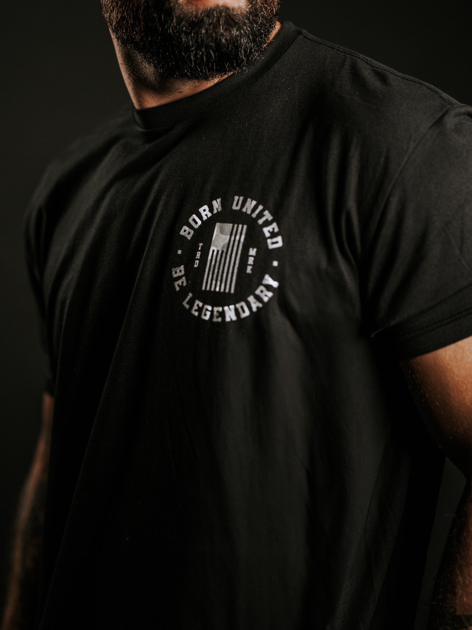 A bearded man wears a black Legend Dynasty t-shirt by Born United Private Label, featuring a white chest emblem with BORN UNITED and BE LEGENDARY encircling a stylized flag. The background is dark.