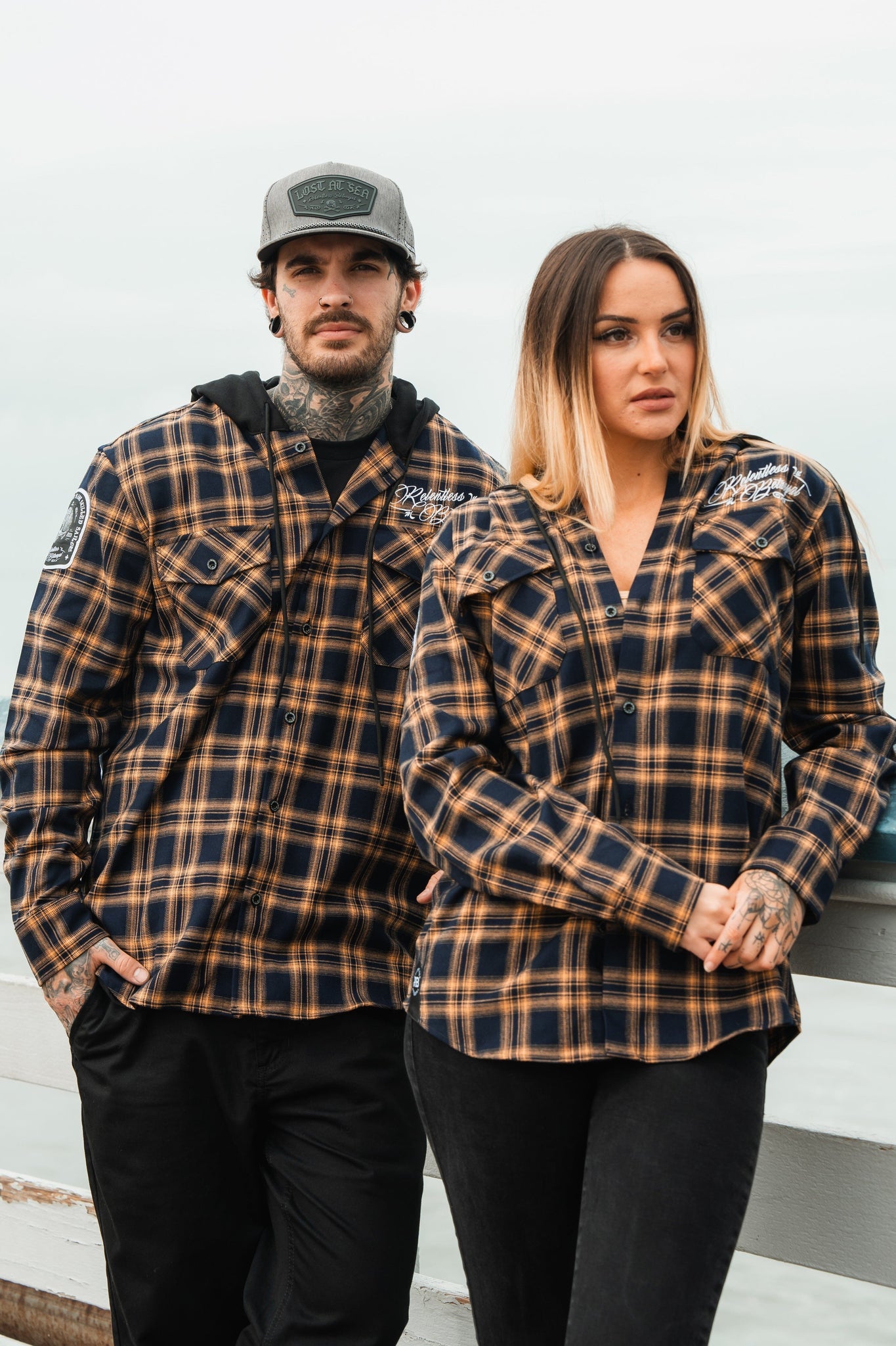 A man and woman in matching Relentless Betrayals Skilled Sailor Premium Hooded Flannels and dark pants stand side by side. The man, wearing a cap, has his arm on a railing as the woman stands with her hands by her sides under a pale sky outdoors.