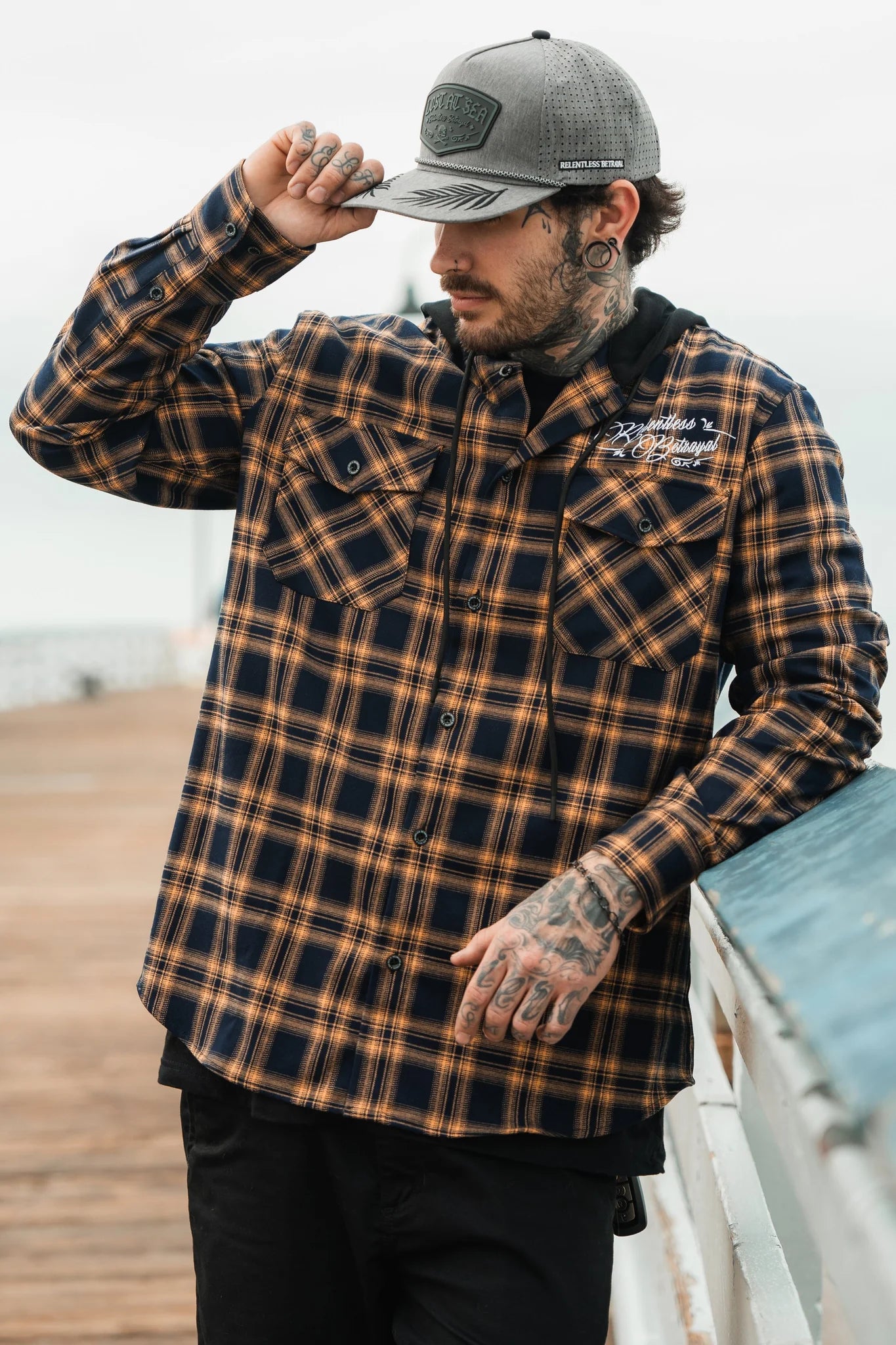 A person in a gray cap and a plaid shirt from Relentless Betrayals Skilled Sailor Premium Hooded Flannel leans on a wooden railing. Outside on a boardwalk, they tilt their cap with one hand, showcasing tattoos on their hand and neck.