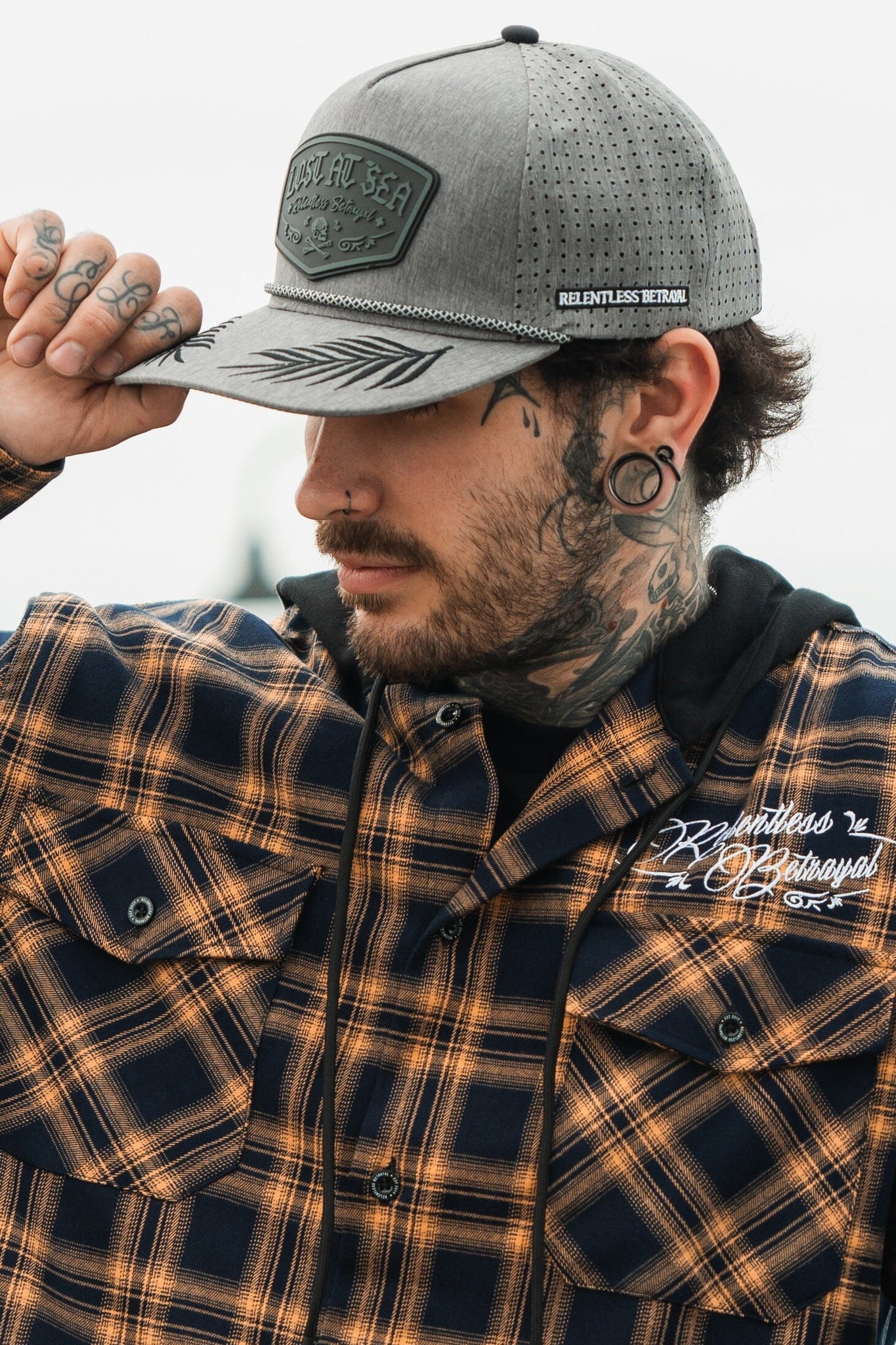 A tattooed man with gauges wears a plaid shirt and the Lost At Sea Gunmetal PVC Snapback by Relentless Betrayal. Outdoors, he adjusts the water-resistant cap with one hand, displaying finger tattoos, while a blurred background completes the scene.