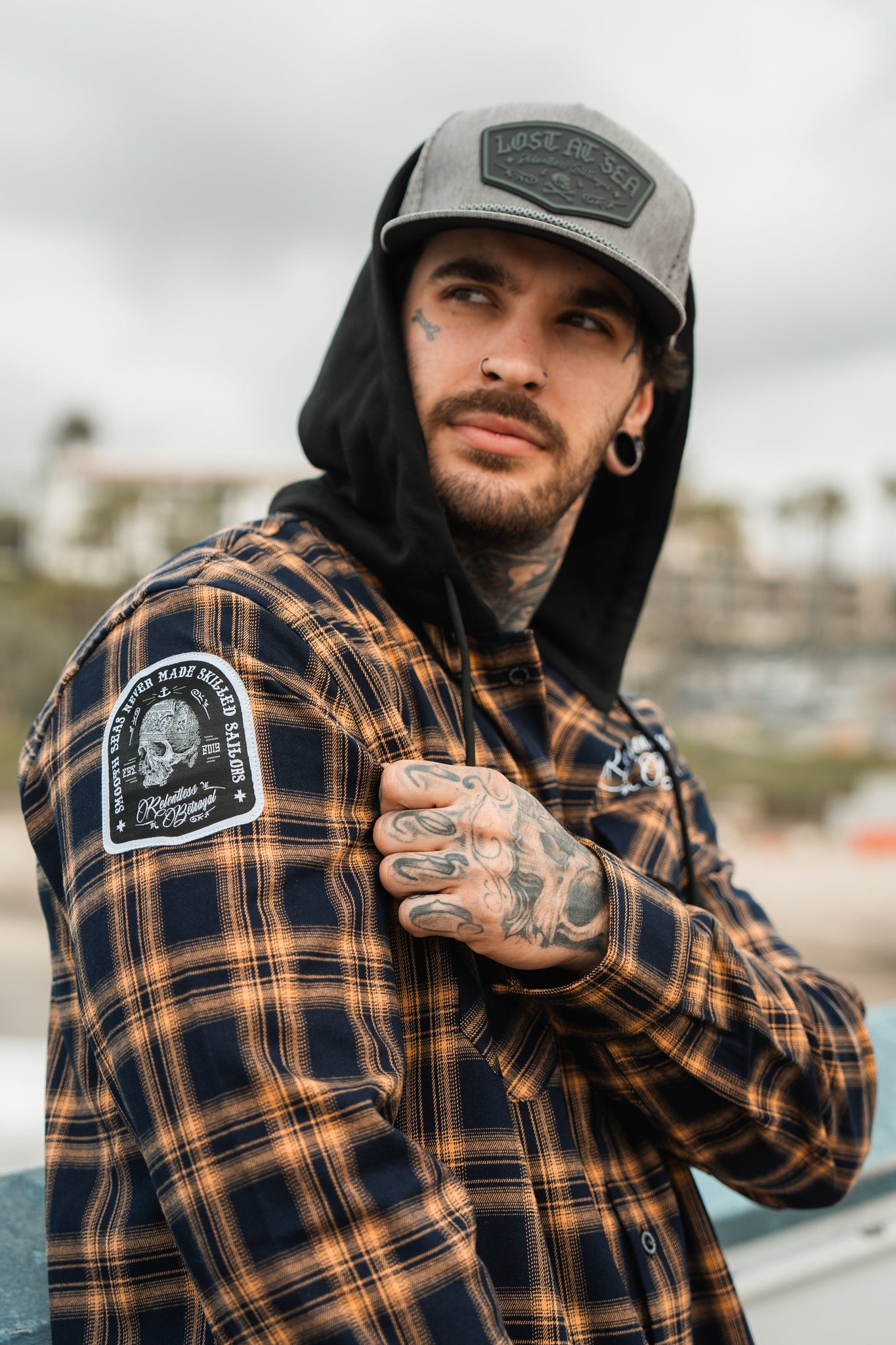 A tattooed individual, wearing an ultra-soft flannel checkered shirt, a Relentless Betrayal Skilled Sailor Premium Hooded Flannel, and a cap, gazes sideways. Their cotton/polyester blend outfit contrasts with the blurred outdoor background.
